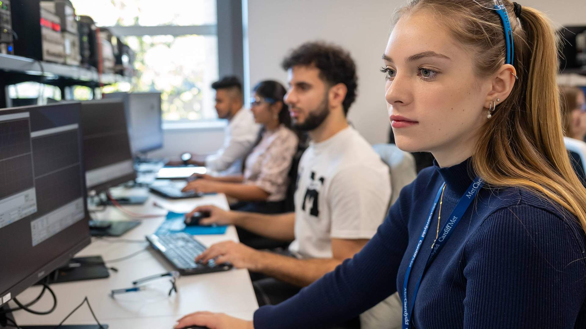 A group of people sit alongside each other at a long desk. Each of them is using a computer workstation.