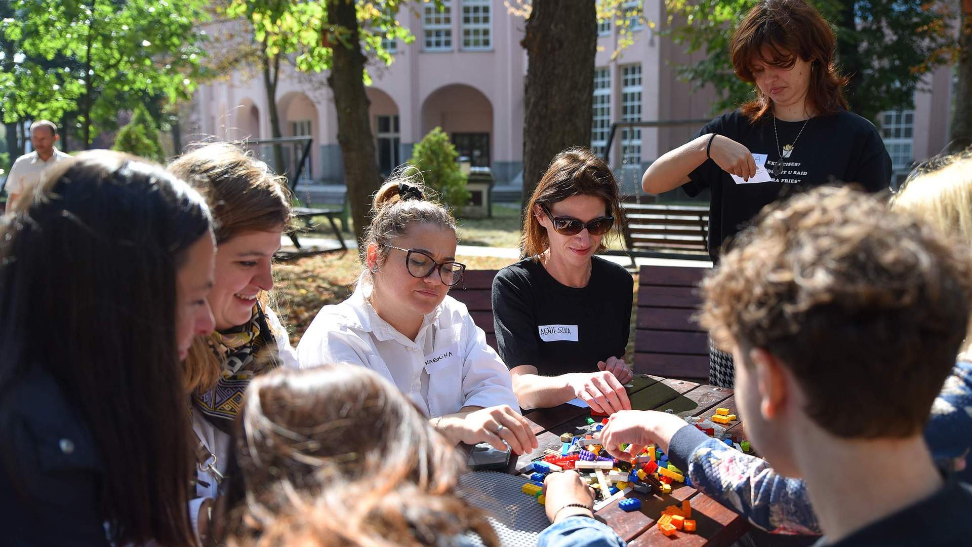 A group of people wearing name badges sit around a table outdoors. On the table in front of them are LEGO bricks.