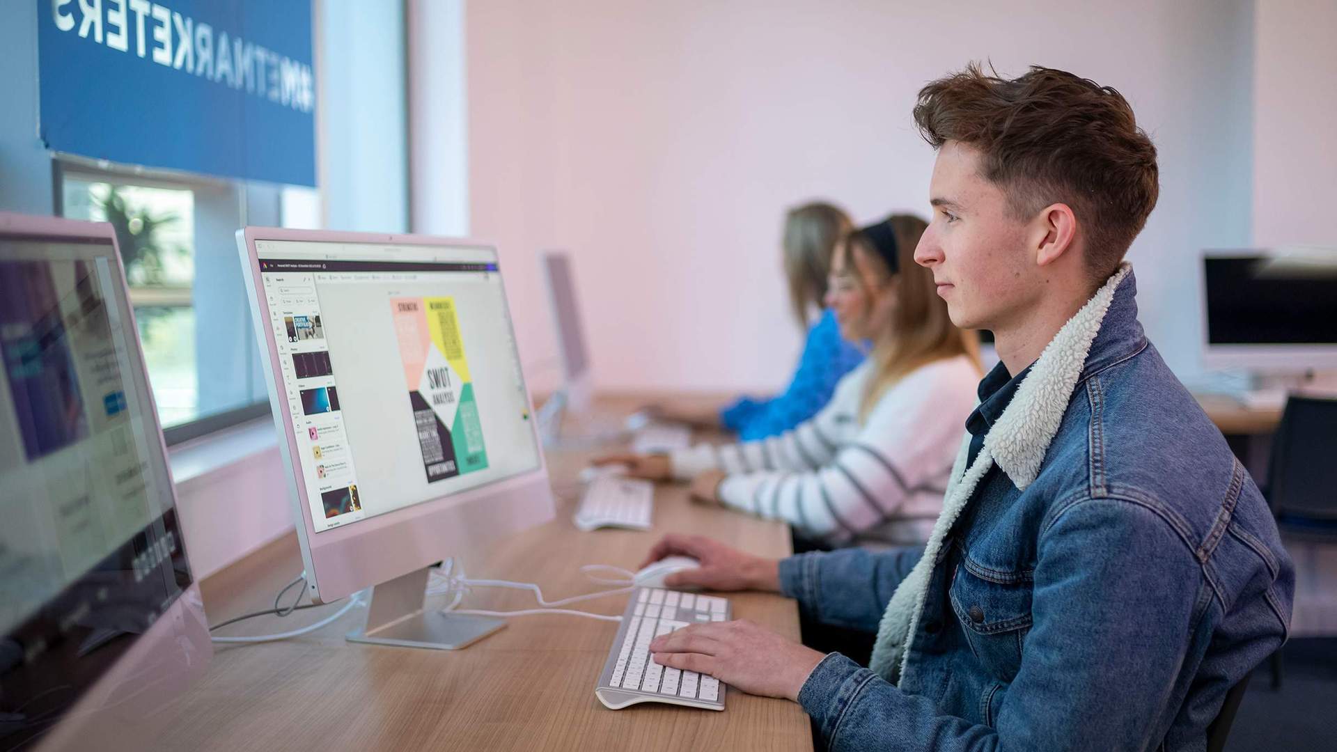 A person sat at a desk in front of a Mac computer. In the background are two other people also using computers.