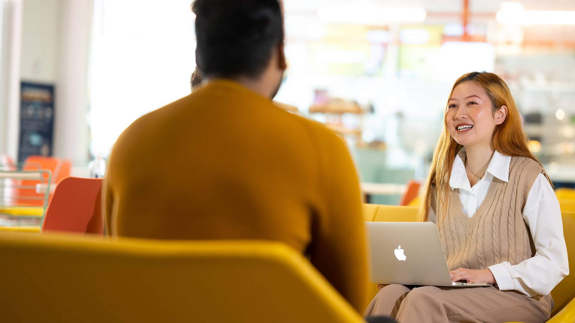 A young woman sits on a yellow chair facing another person. A Mac laptop computer is resting on her lap.