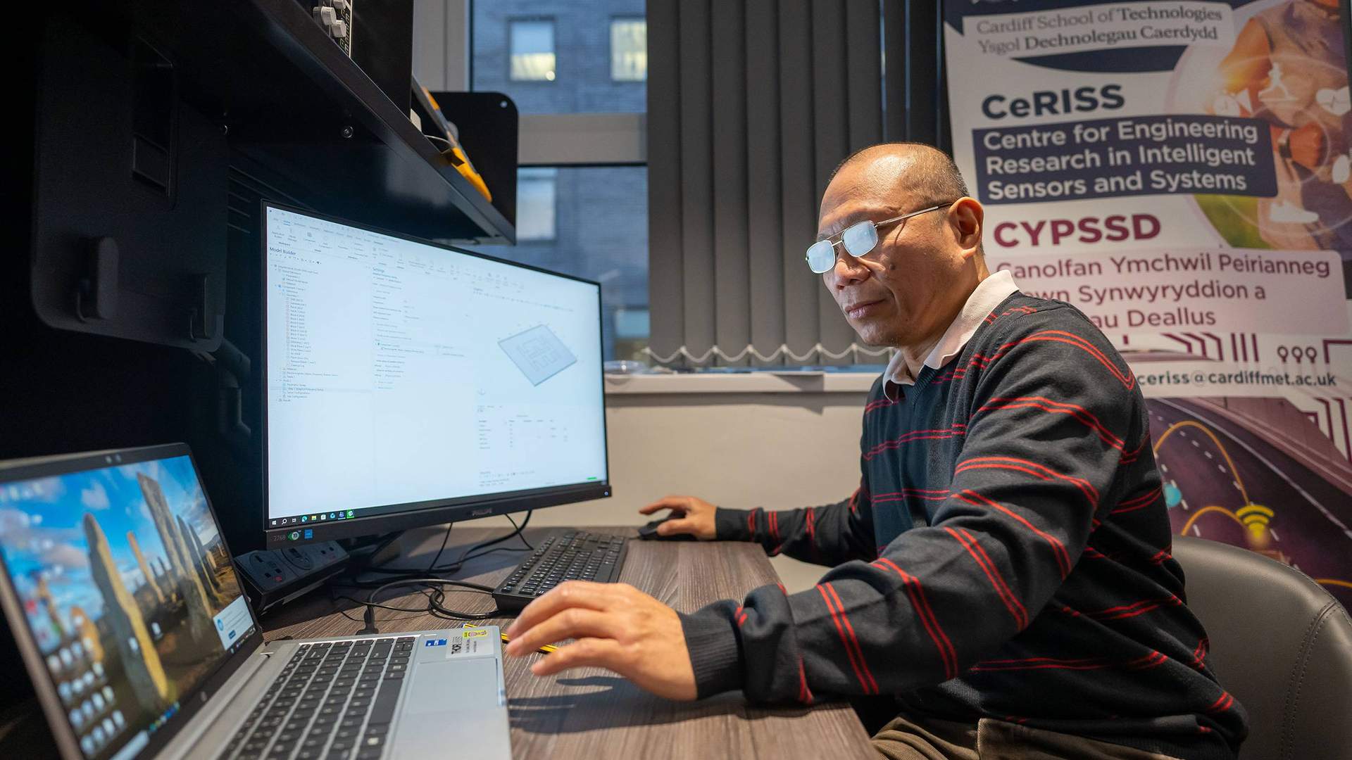 A person sitting at a desk uses a computer workstation. Their right hand controls the mouse, while their left reaches for the trackpad of an open laptop computer.