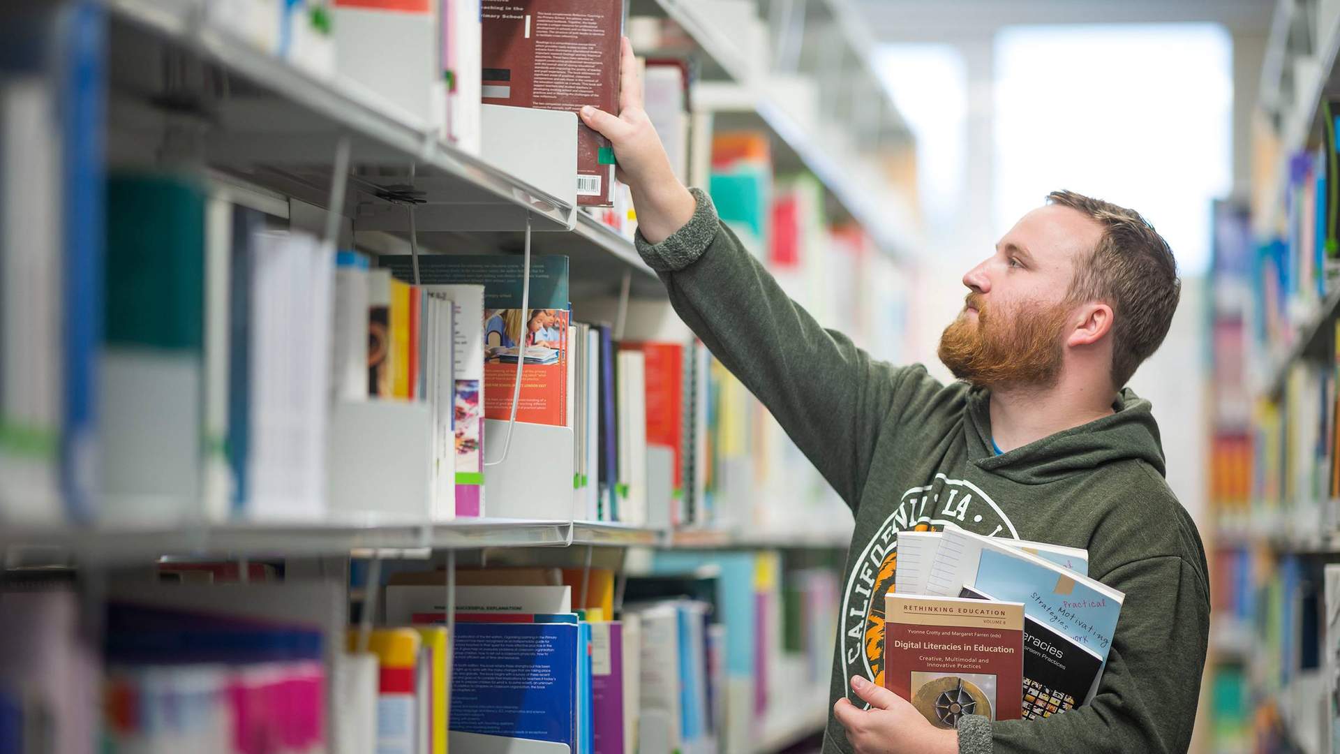 A young man with a beard reaches for a book on a library shelf, while holding several other books underneath his arm.