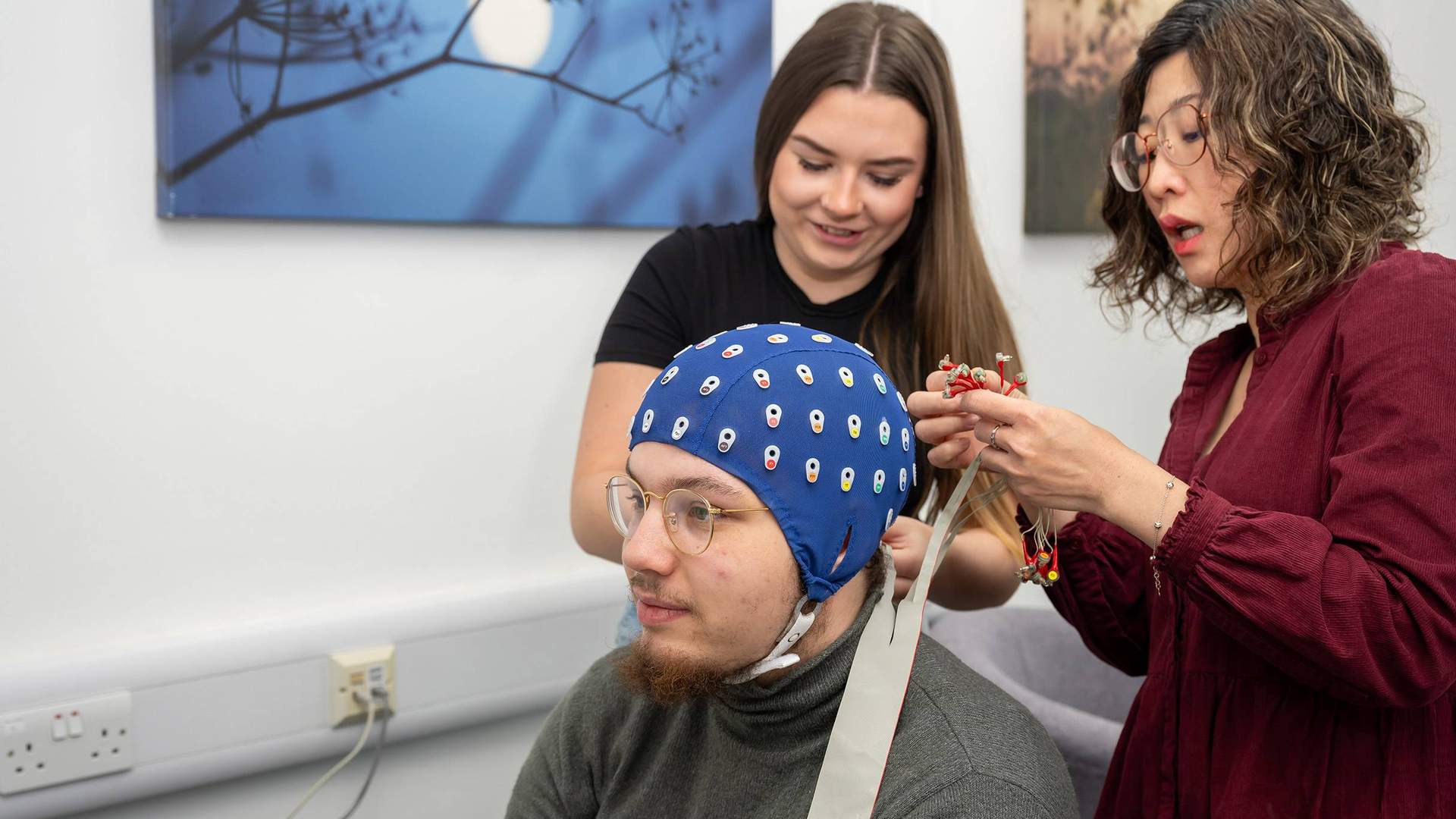 Two people assist a seated person with an EEG cap. One adjusts the cables on the cap, while the other looks on.
