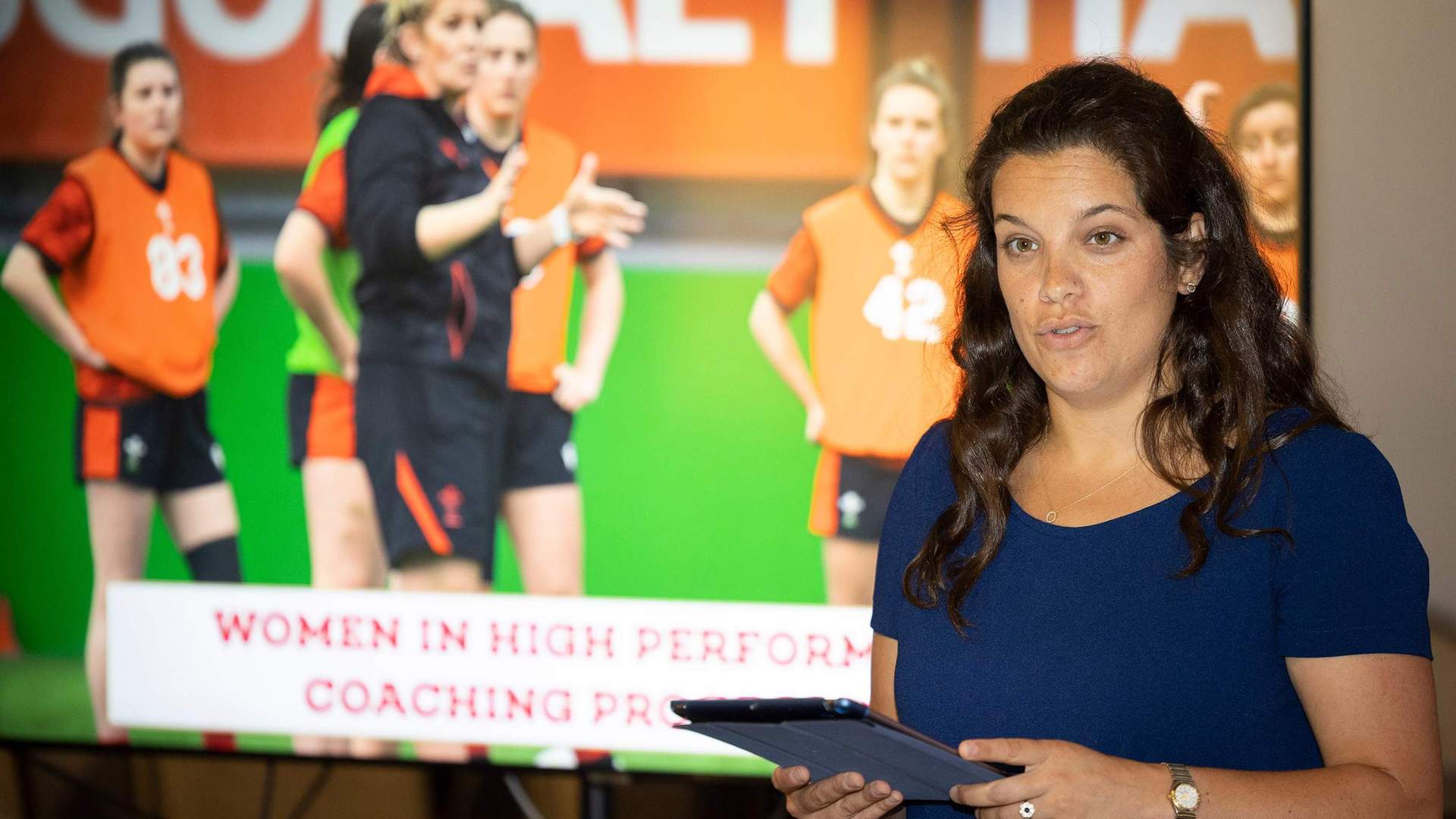 A woman holding an iPad stands in front of large display screen. On the screen are a group of women's rugby players.