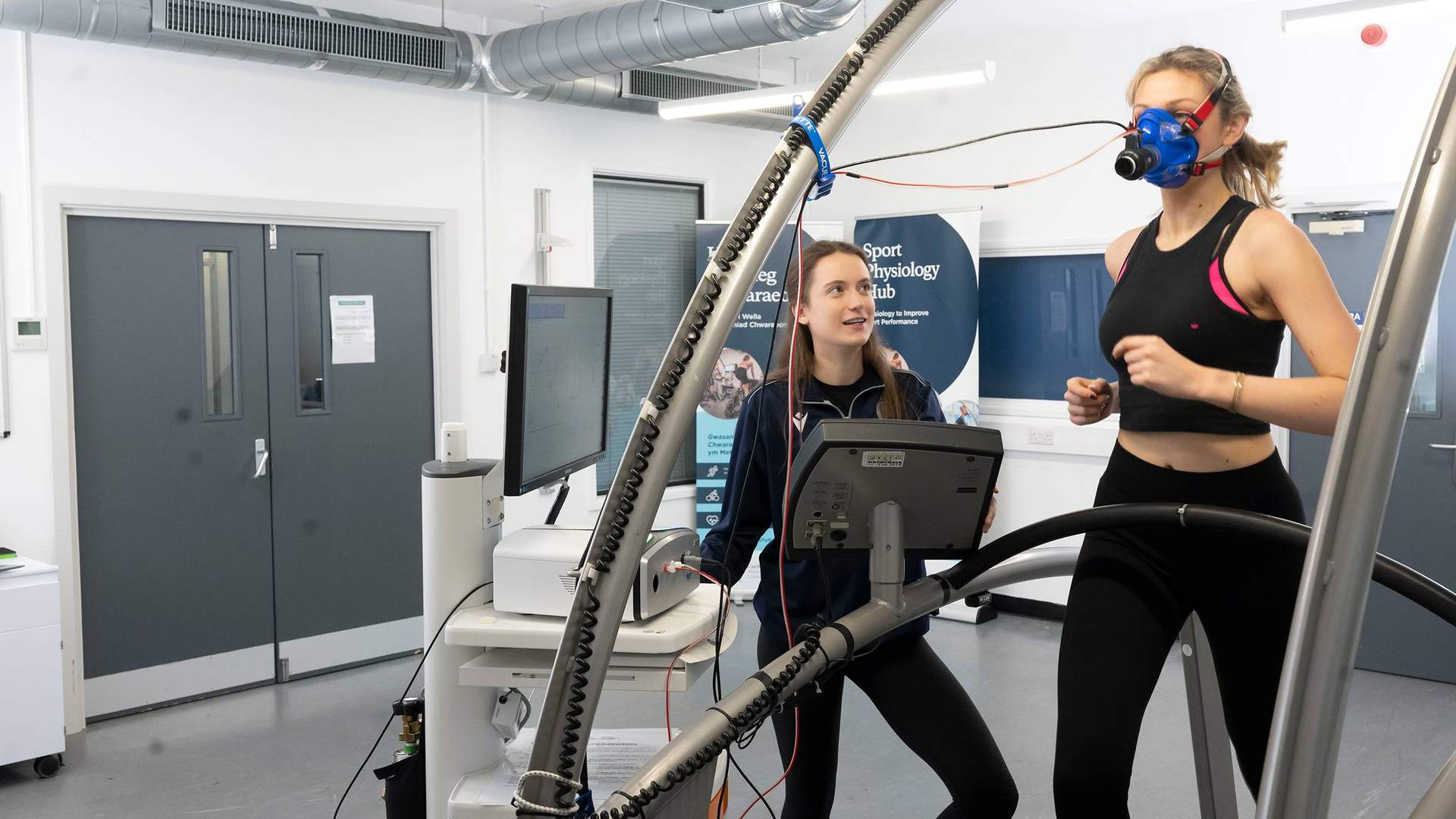 A person runs on a treadmill while wearing a breathing mask. A second person stands alongside, next to a computer display and other equipment.