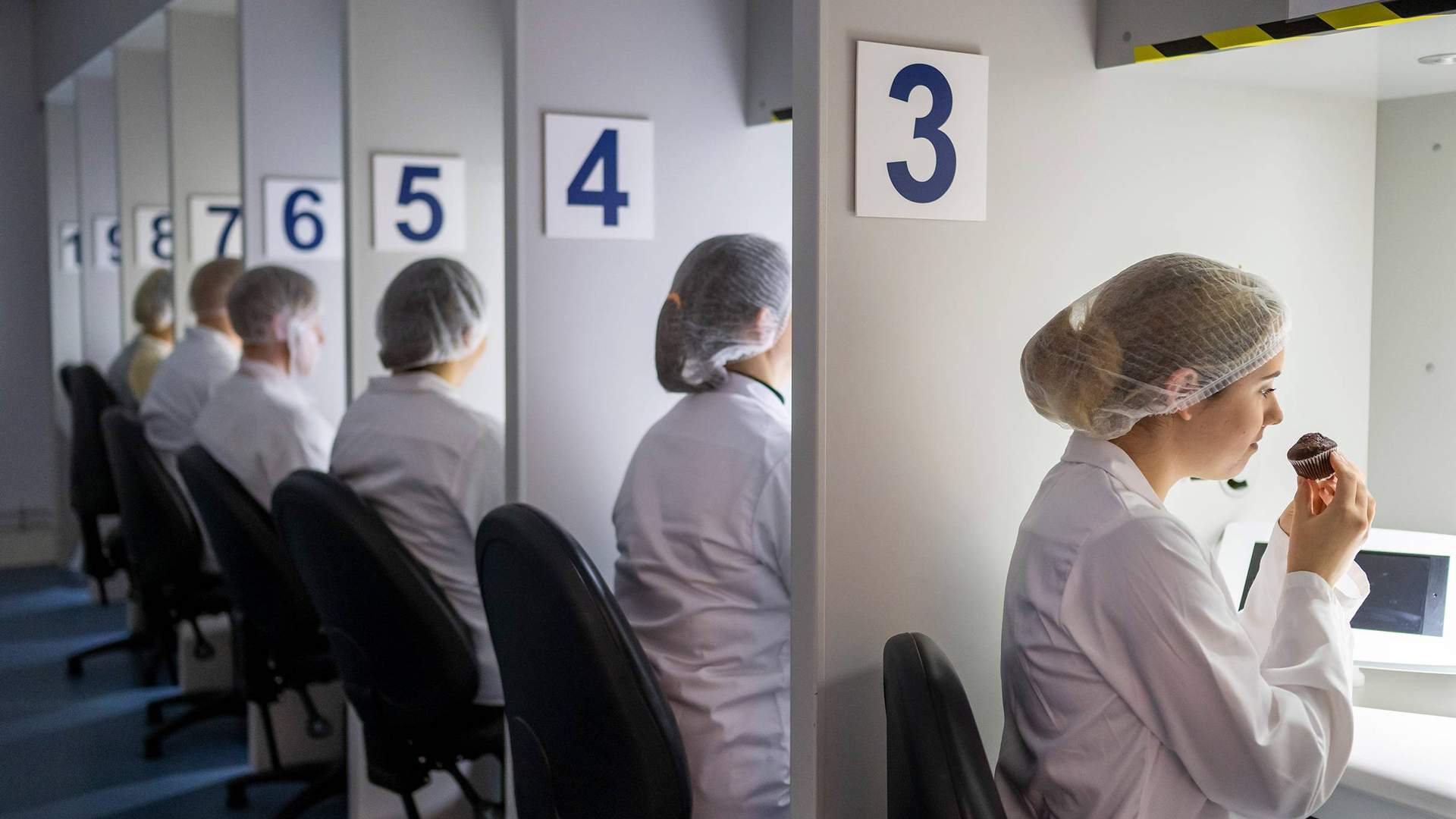 A row of people in white lab coats and hairnets sit at numbered stations. Each person is engaging in a sensory evaluation, with one person smelling or tasting from a small cake under a hooded light. The stations are numbered 3 to 8.