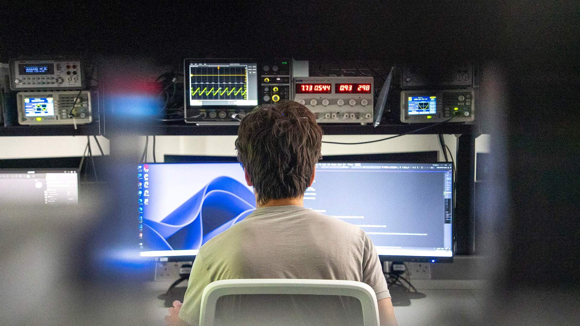 Student facing two computer monitors and a rack of electronic equipment
