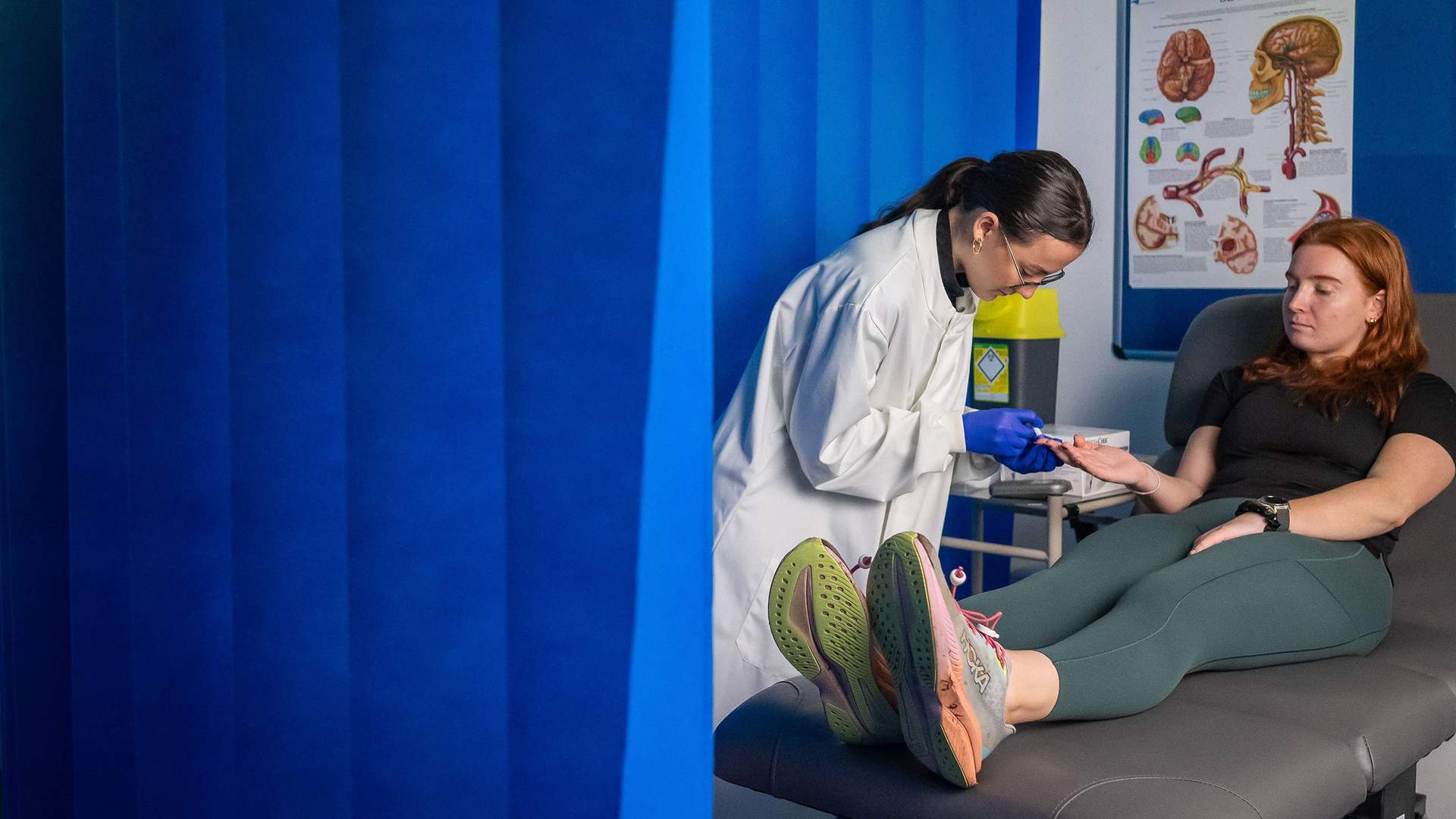 A person in a white lab coat takes a blood sample from the finger of a patient, sat on an examination bed. They are surrounded by a blue floor-to-ceiling curtain.