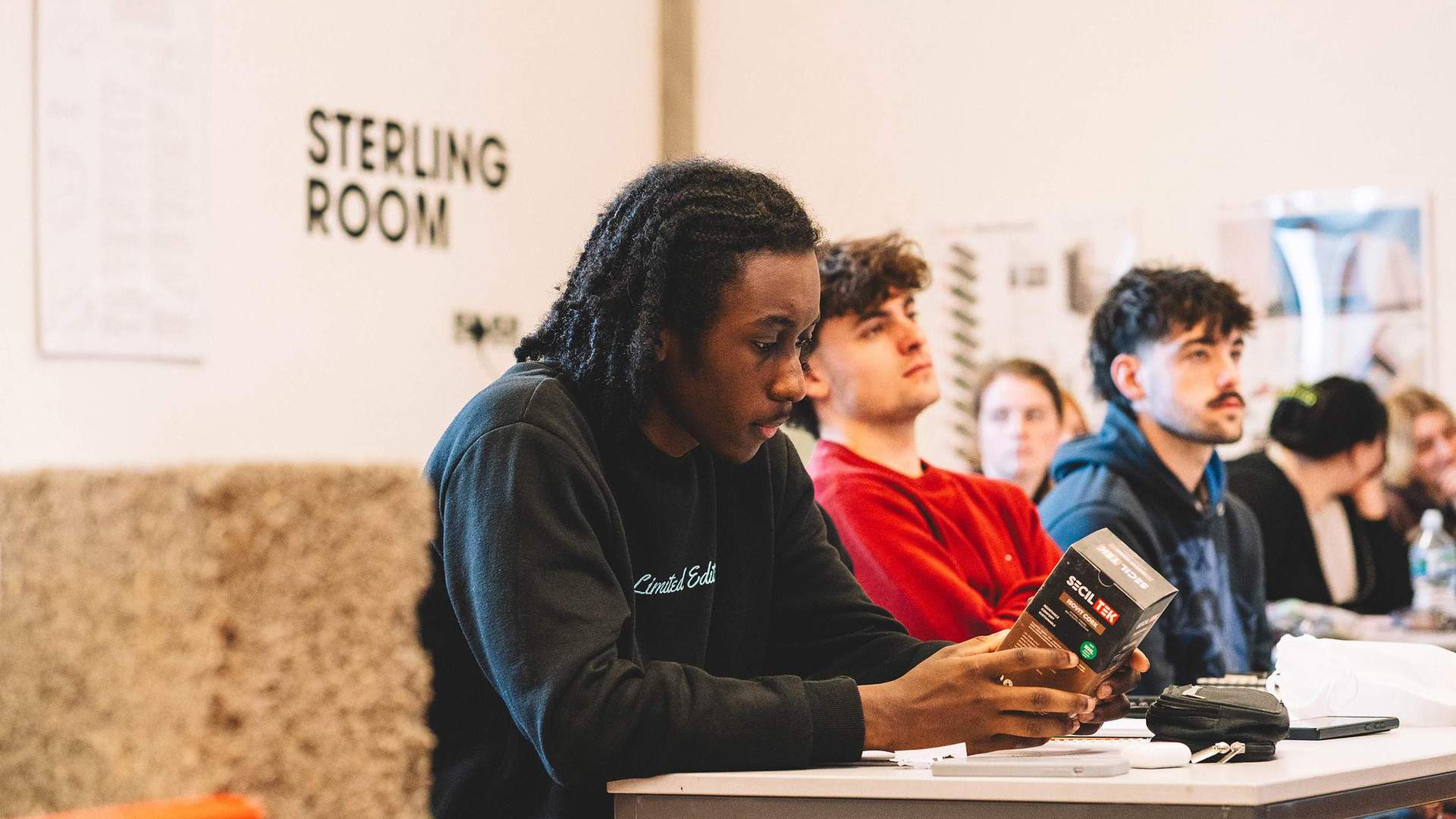 Student holding a boxed product while seated at a classroom table with others listening nearby.