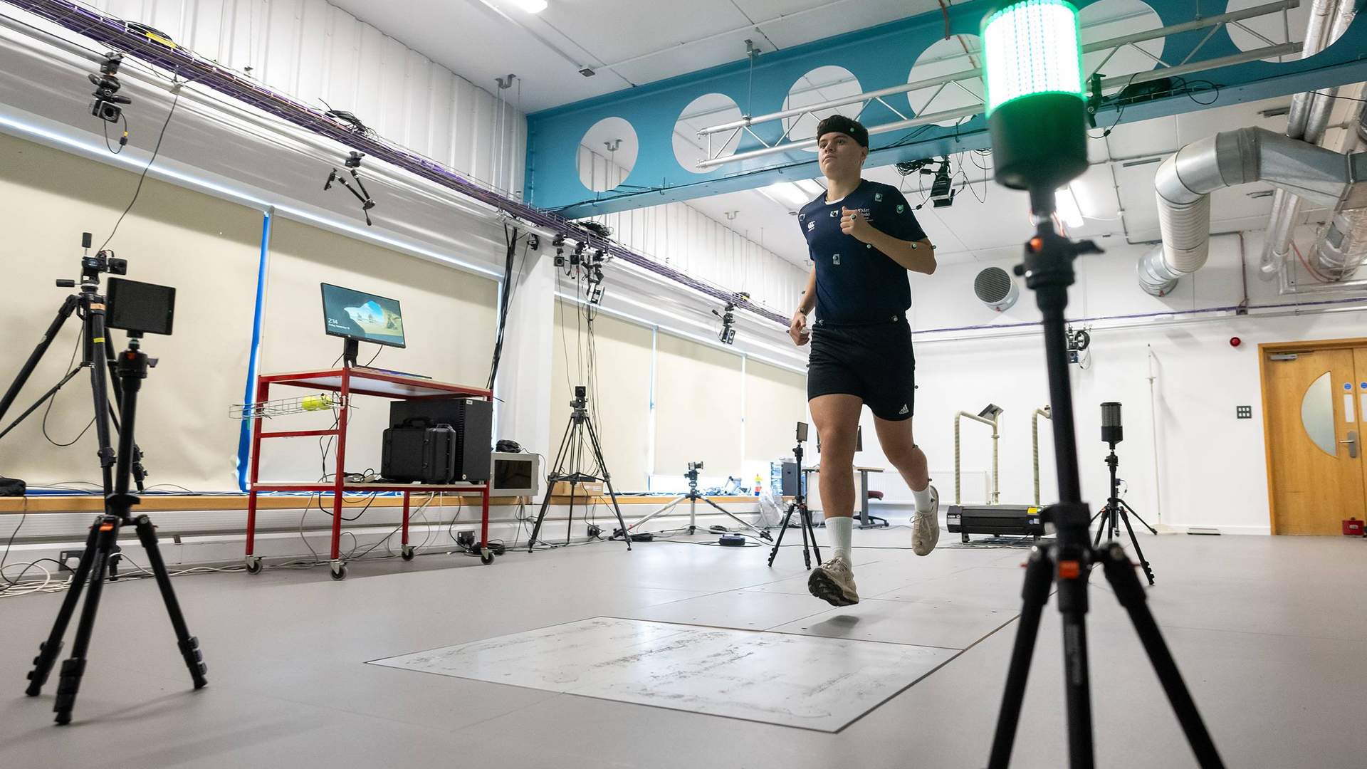 A person runs in a technology-equipped lab, surrounded by cameras and a computer screen. A bright green light is visible in the foreground. The setting suggests a motion analysis or biomechanics study.
