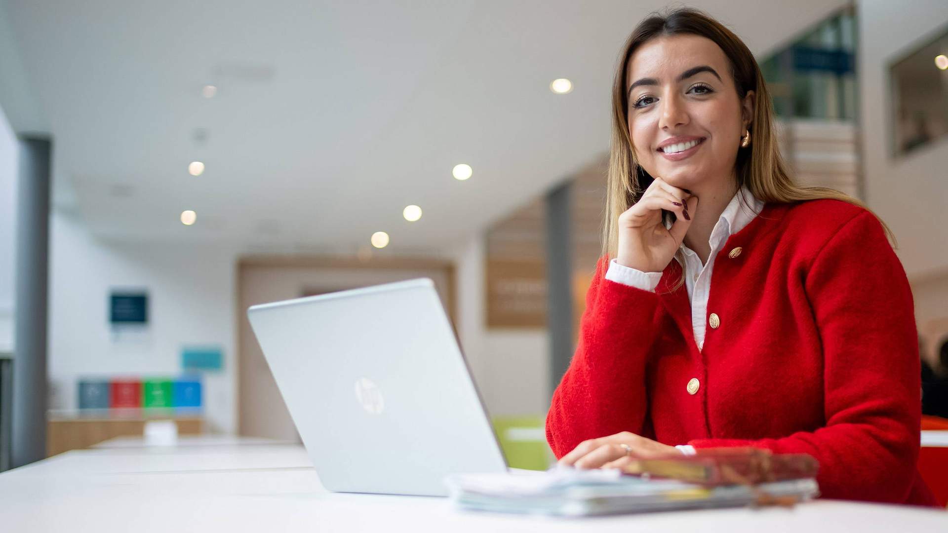 A person is smiling while sitting at a table with a laptop in front of them. They are in a bright, modern setting.