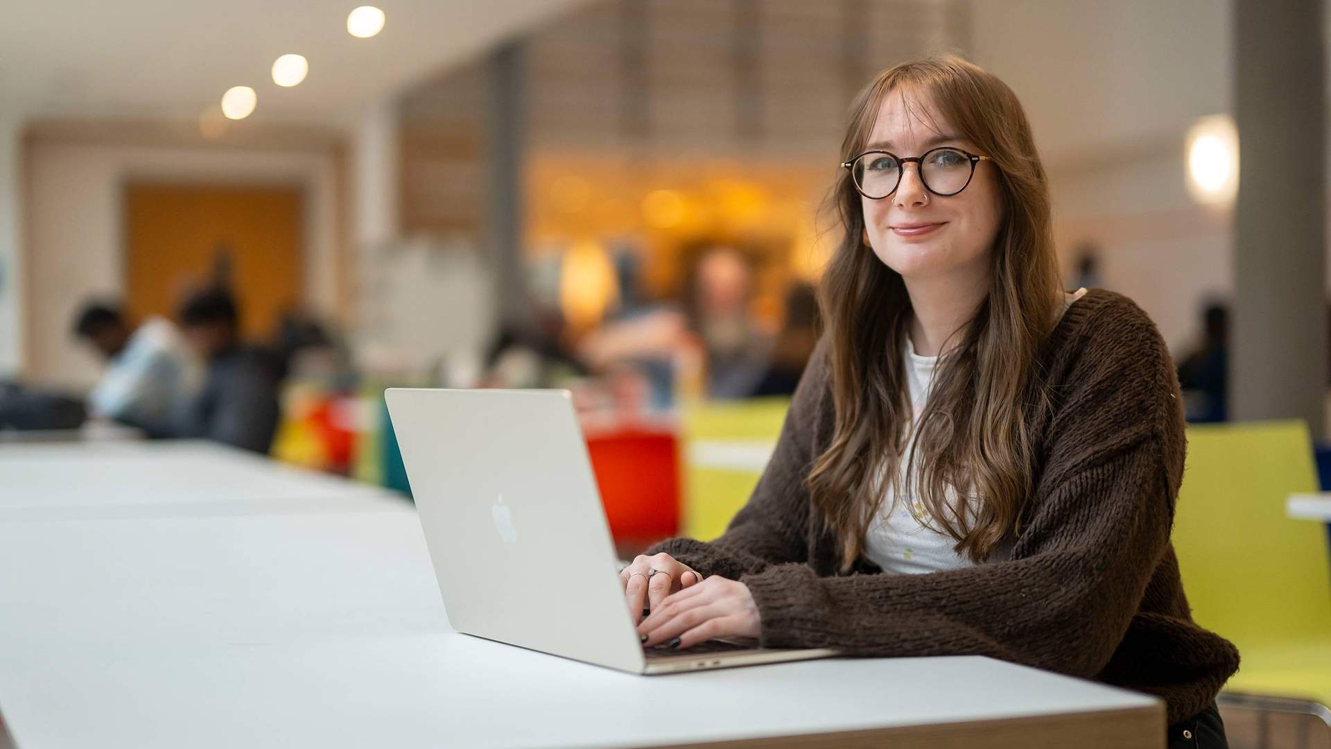 A person is smiling while sitting at a table with a laptop in front of them. They are in a bright, modern setting.