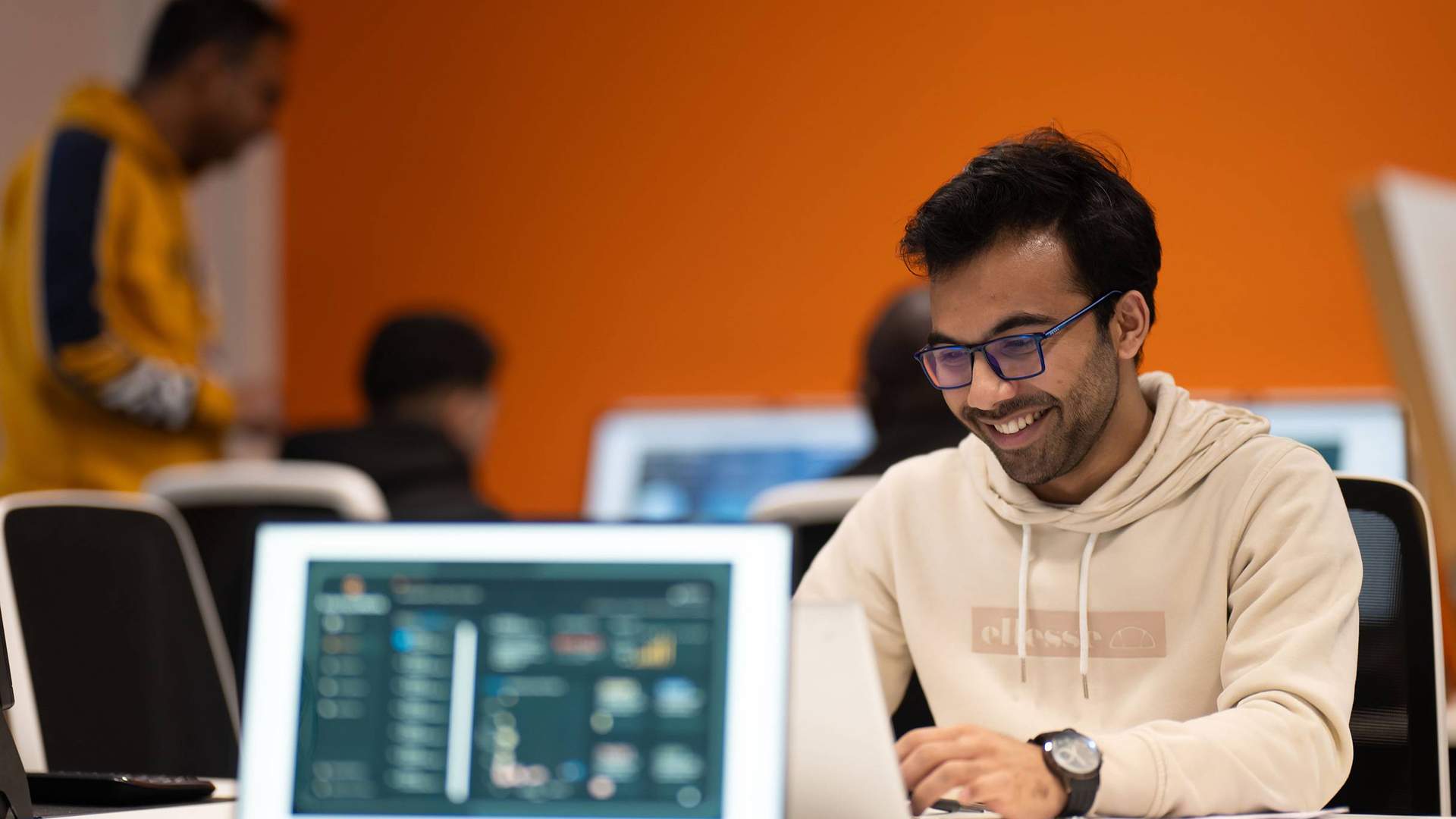 A student wearing a white hoodie and glasses is focused on his laptop while working at a desk.