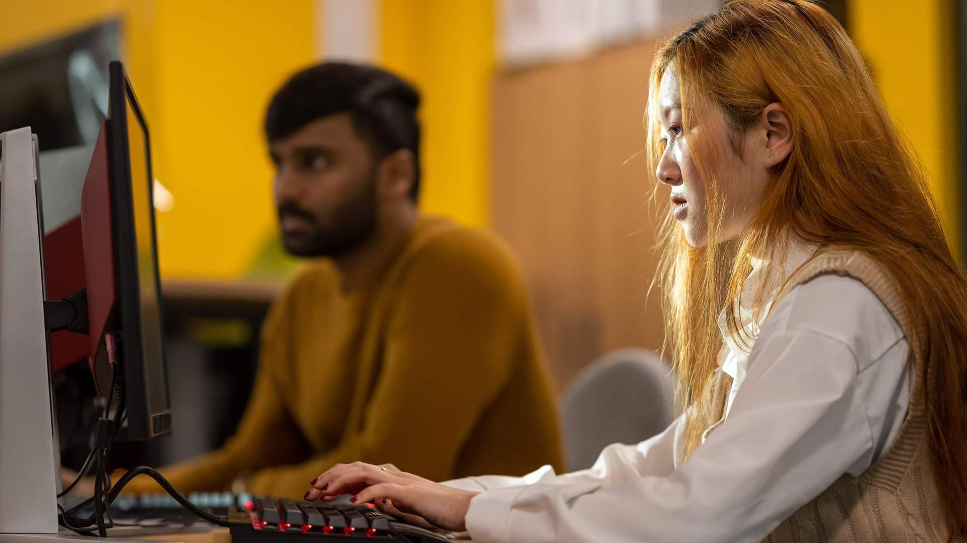 A person working intently at a computer workstation. In the background another person also works at a computer.