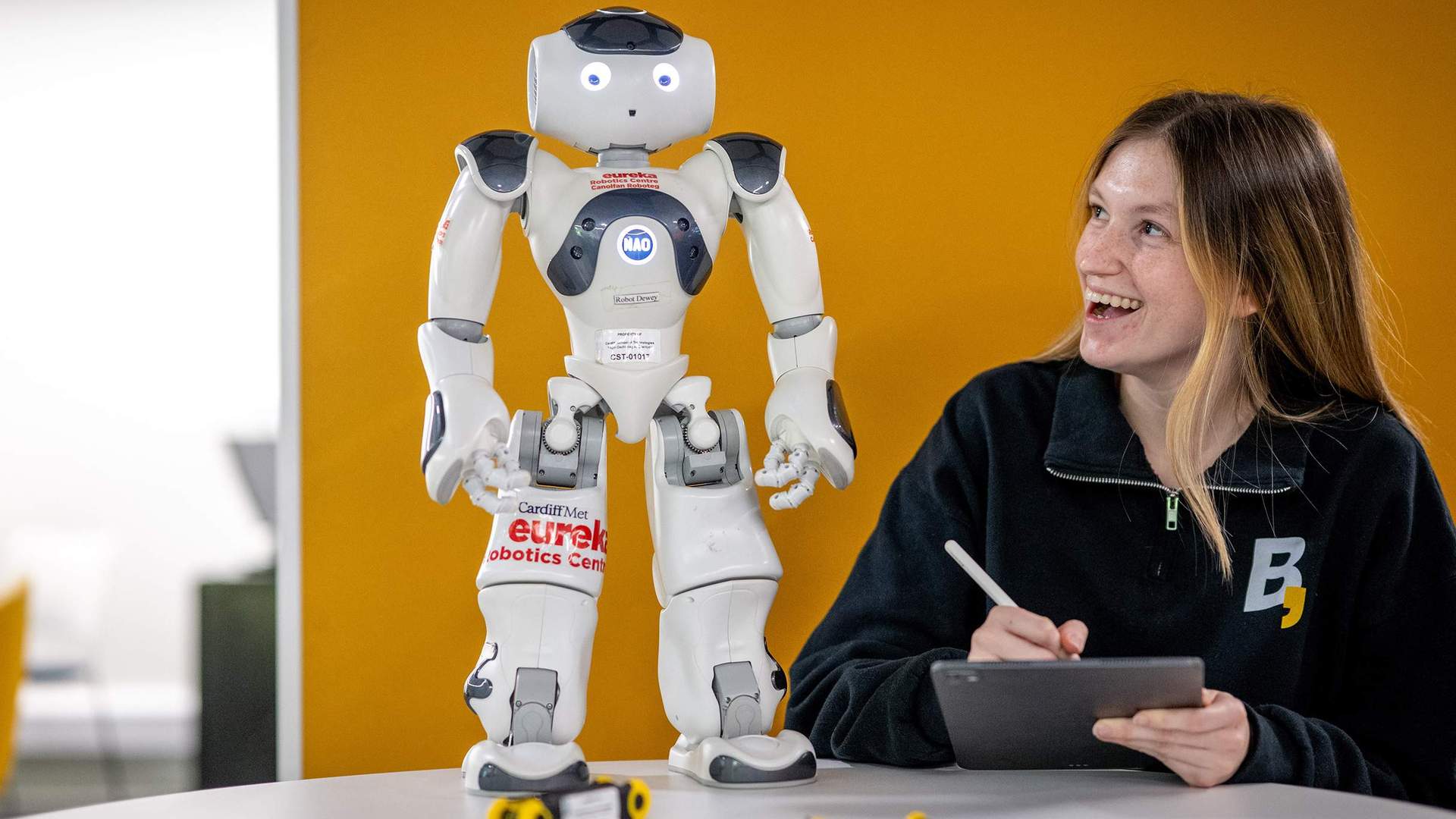 Person holding a tablet and stylus while looking up at a small humanoid robot.