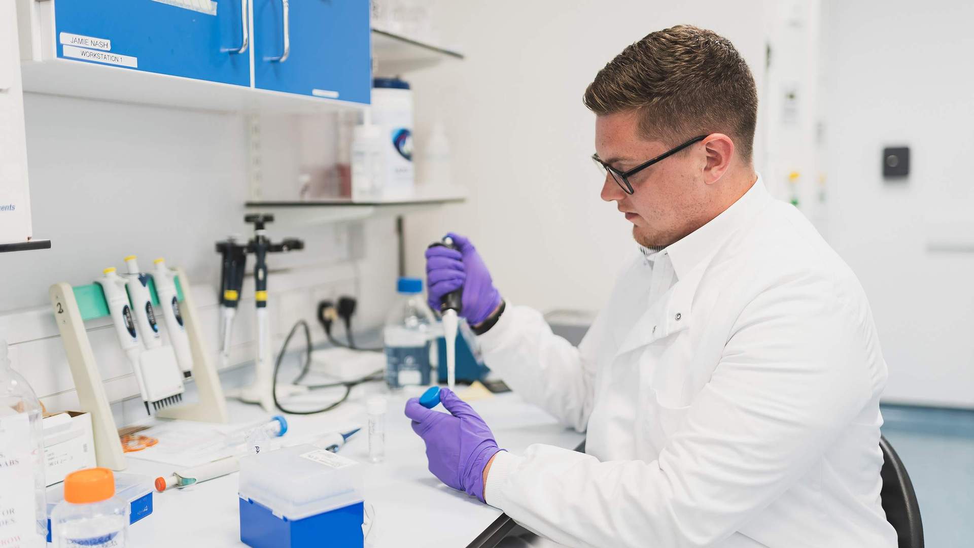 A person in a lab coat uses a pipette to dispense a sample into a container.