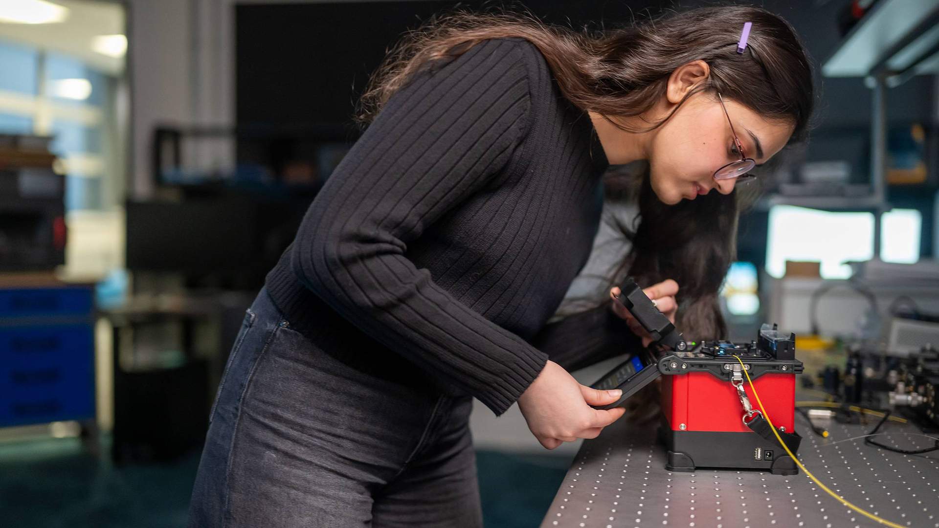 A student pressing the touchscreen of a small electronic device. In the background are other devices and computer monitors.