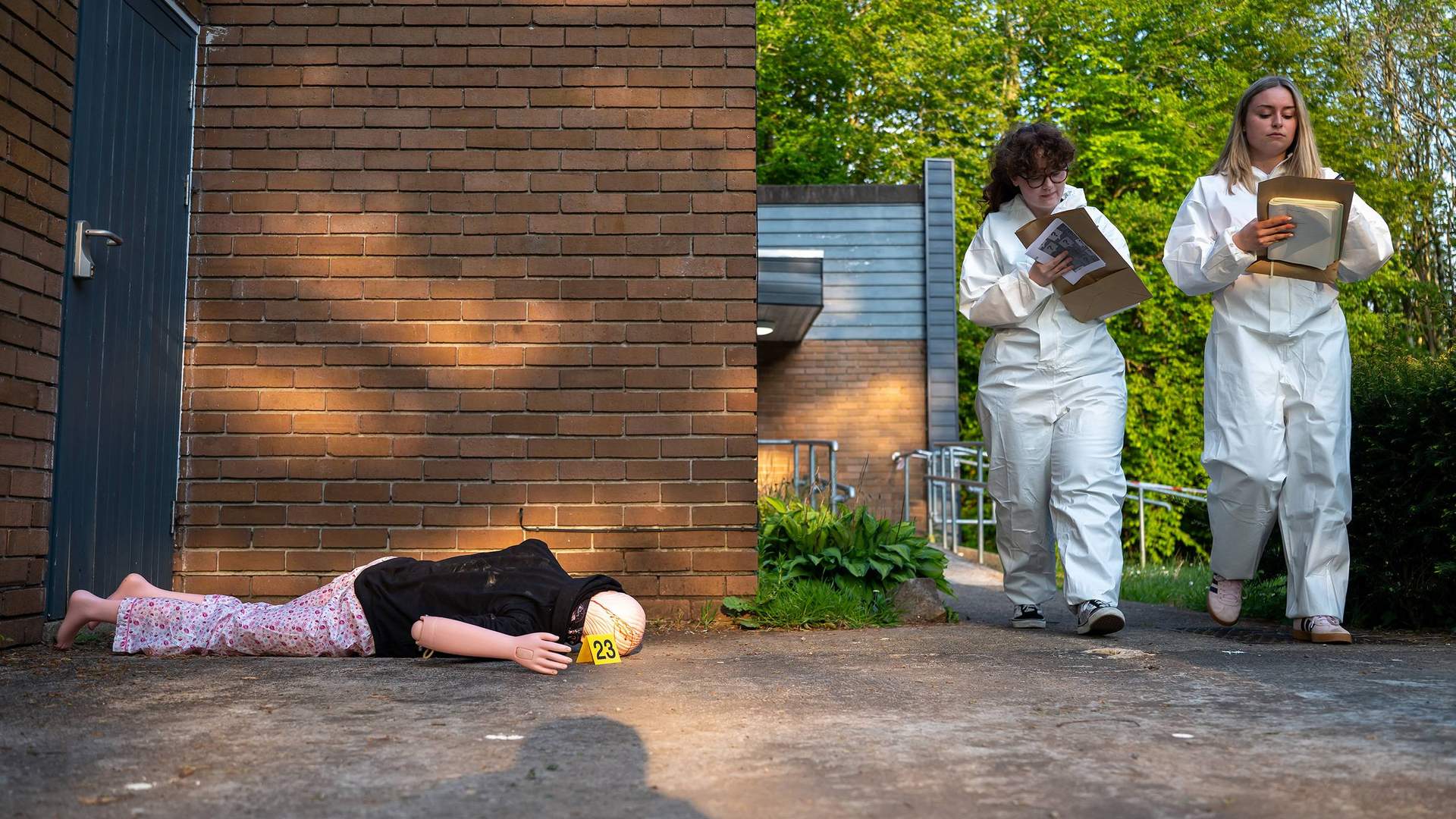 Two students wearing white overalls walk through a simulated crime scene. On the ground is a mannequin with a yellow evidence marker next to it.