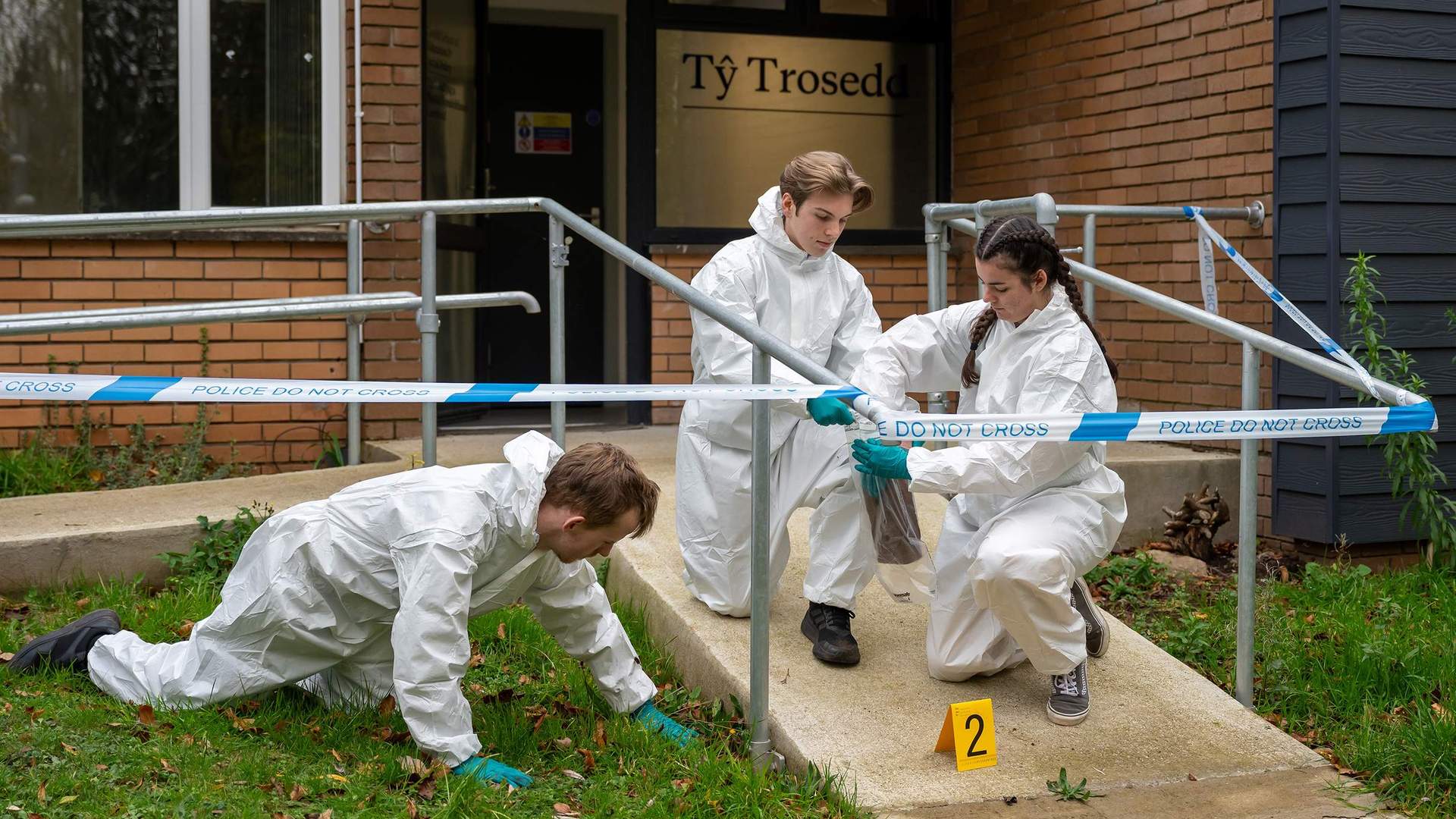 Three students wearing white jumpsuits and gloves inspect a fake crime scene for clues