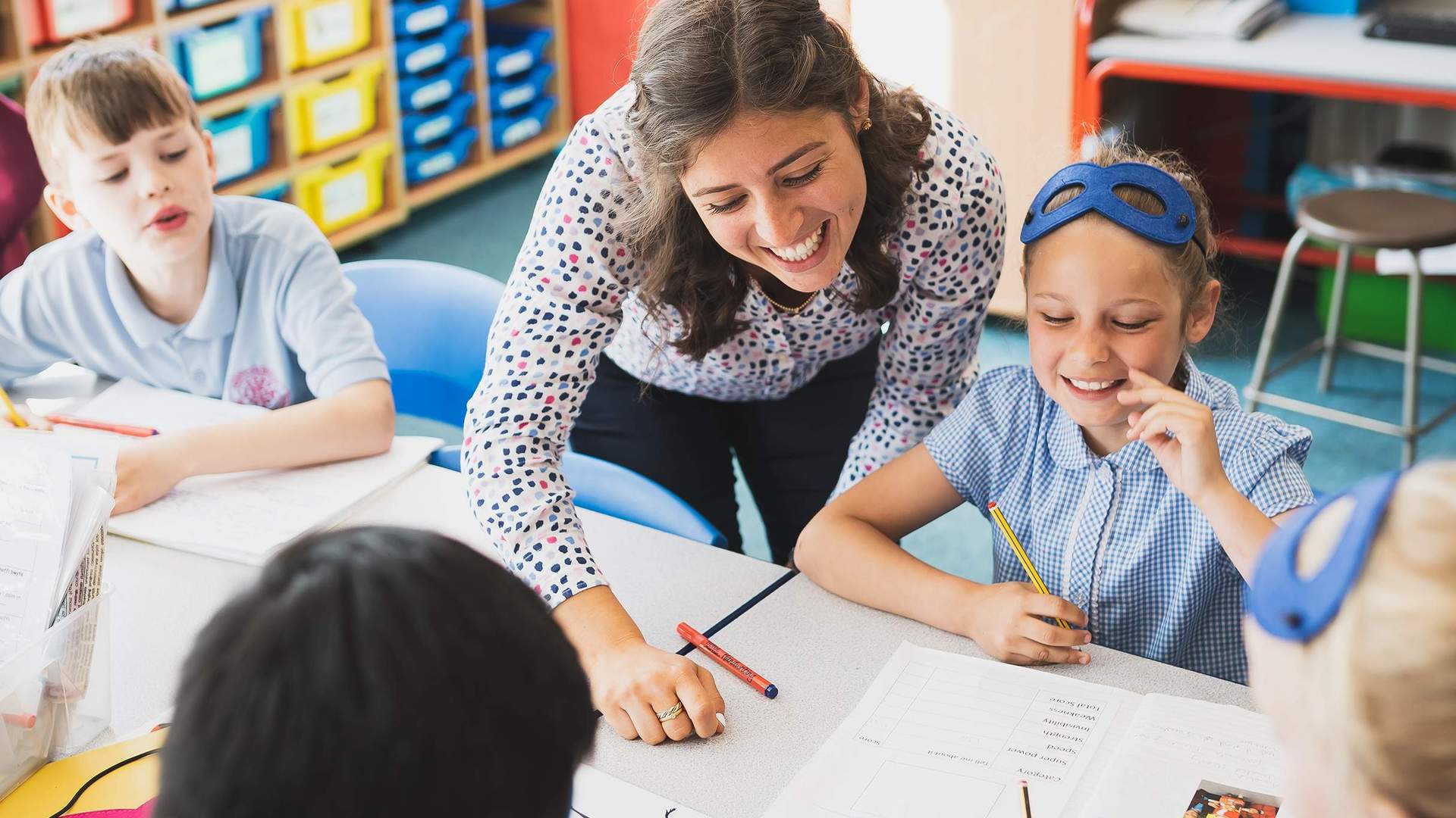 A teacher between two pupils sitting at a desk. The teacher is looking at an open book in front of one of the pupils.