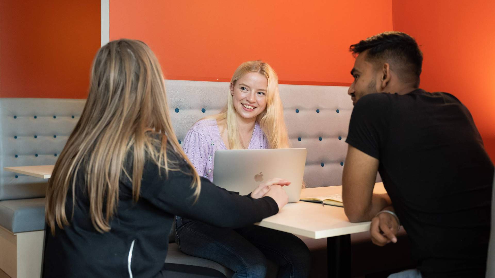 Three students sit together on bench seating at a table. The wall behind them is a vibrant orange red.