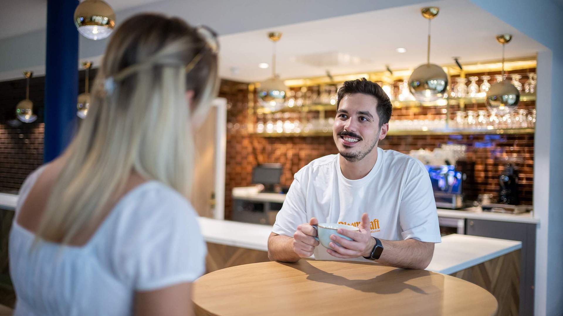 Two people sit at a table talking to each other. In the background is a bar area with a coffee machine.