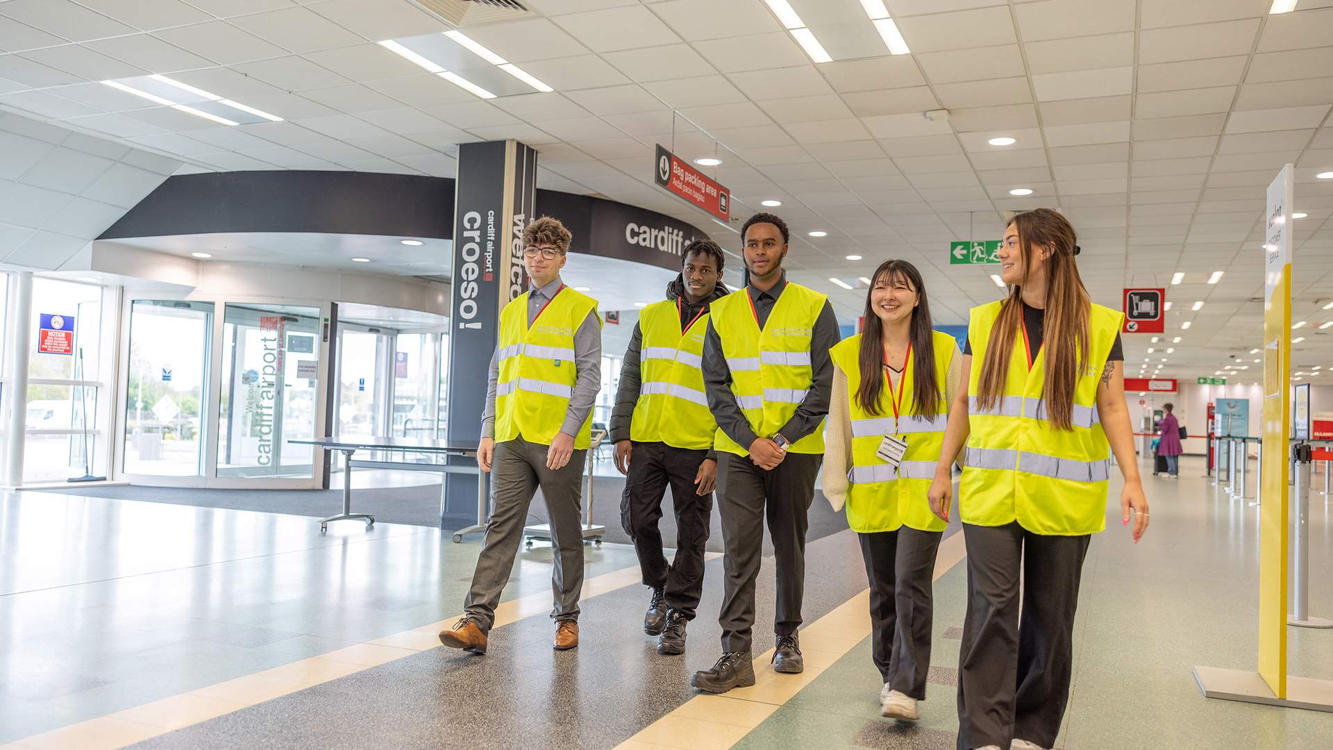 A group of five people wearing yellow safety vests walk through a corridor in an airport. They appear to be conducting an inspection or tour. The corridor is well-lit and has various signs and a glass entrance visible in the background.