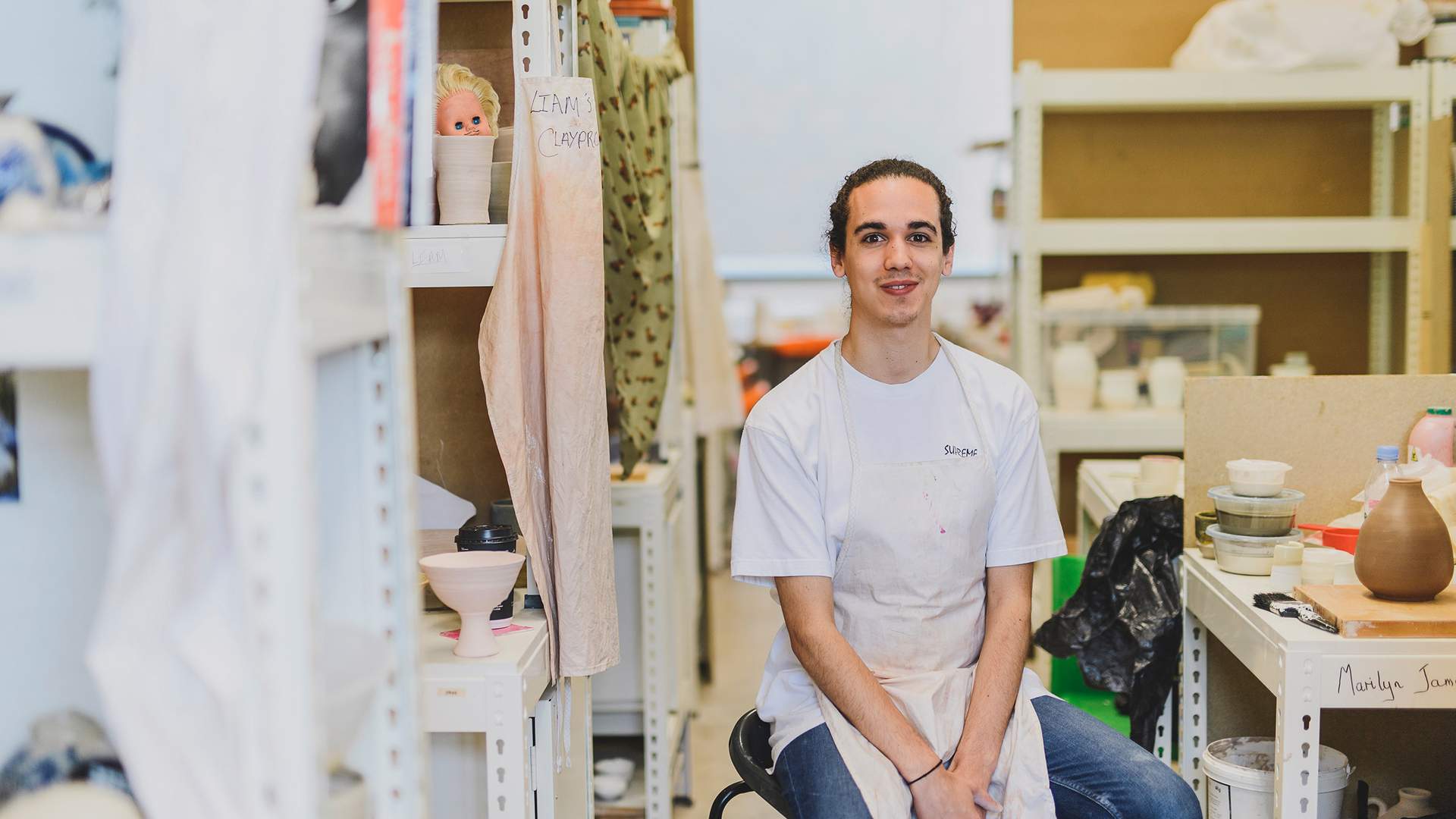 Person with long hair wearing a white apron sits on a stool in an art studio. Shelves filled with pottery, tools, and materials surround them. The environment appears creative and well-used.