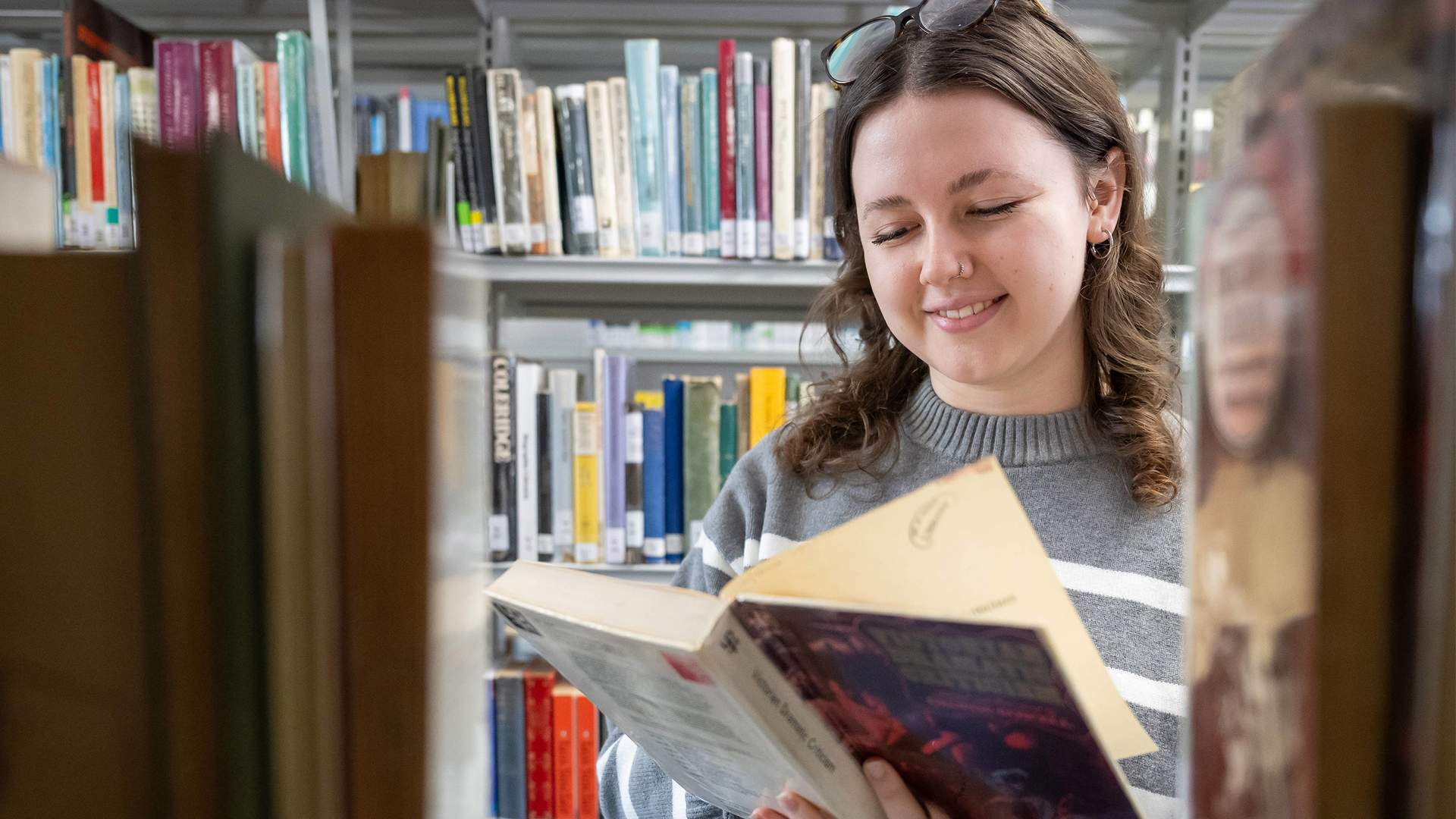 A woman in a striped sweater is smiling while reading a book in a library. She has sunglasses on her head and stands surrounded by shelves filled with books.