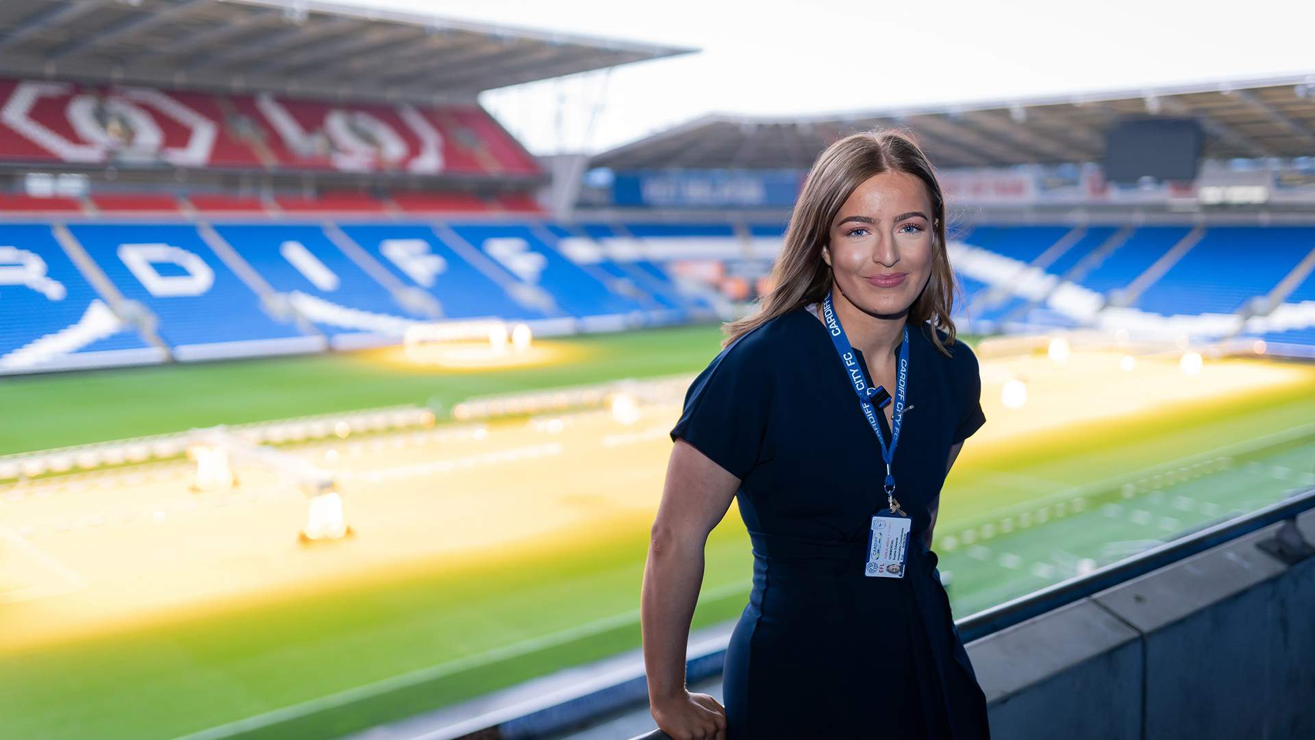 A woman in a navy dress stands smiling on the balcony of an empty stadium. The stadium seats are predominantly blue, with some red sections, and green grass covers the field. She wears an ID lanyard around her neck.