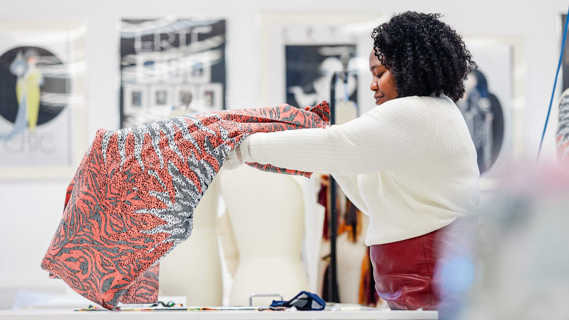 A person with curly hair holds up a colorful fabric in a design studio. In the background, a mannequin and framed art are visible. The room is well-lit, creating an inspiring workspace for creativity.