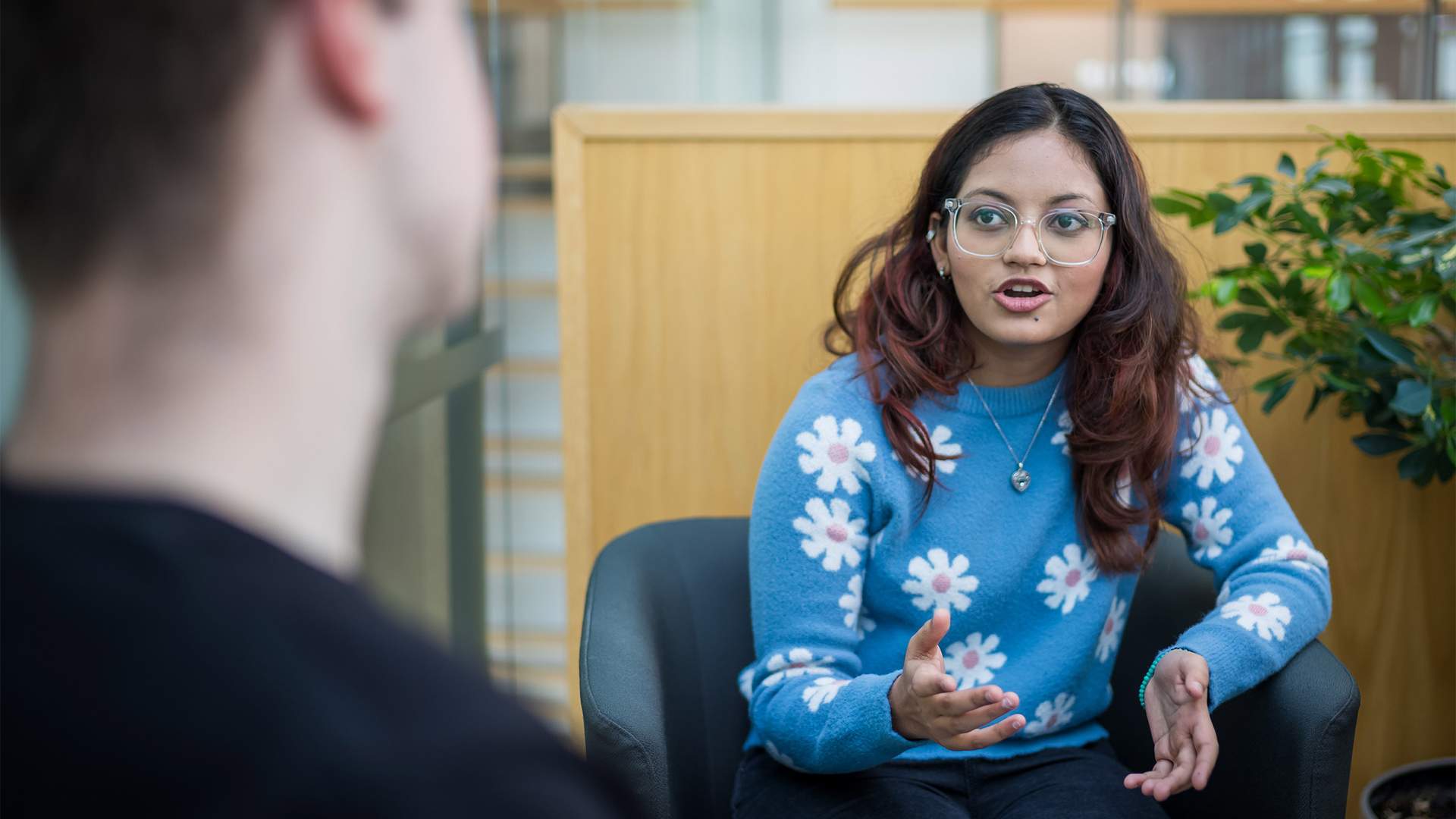 A young woman with glasses and long hair, wearing a blue sweater with white flower patterns, gestures while talking. She is seated in a modern indoor space, facing another person whose back is to the camera. A green plant is in the background.