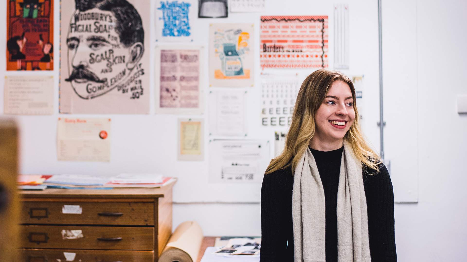 A person with long hair and a scarf stands smiling in an art studio. Behind them is a wall with various vintage posters and prints. A wooden chest of drawers and rolls of paper are visible in the foreground.
