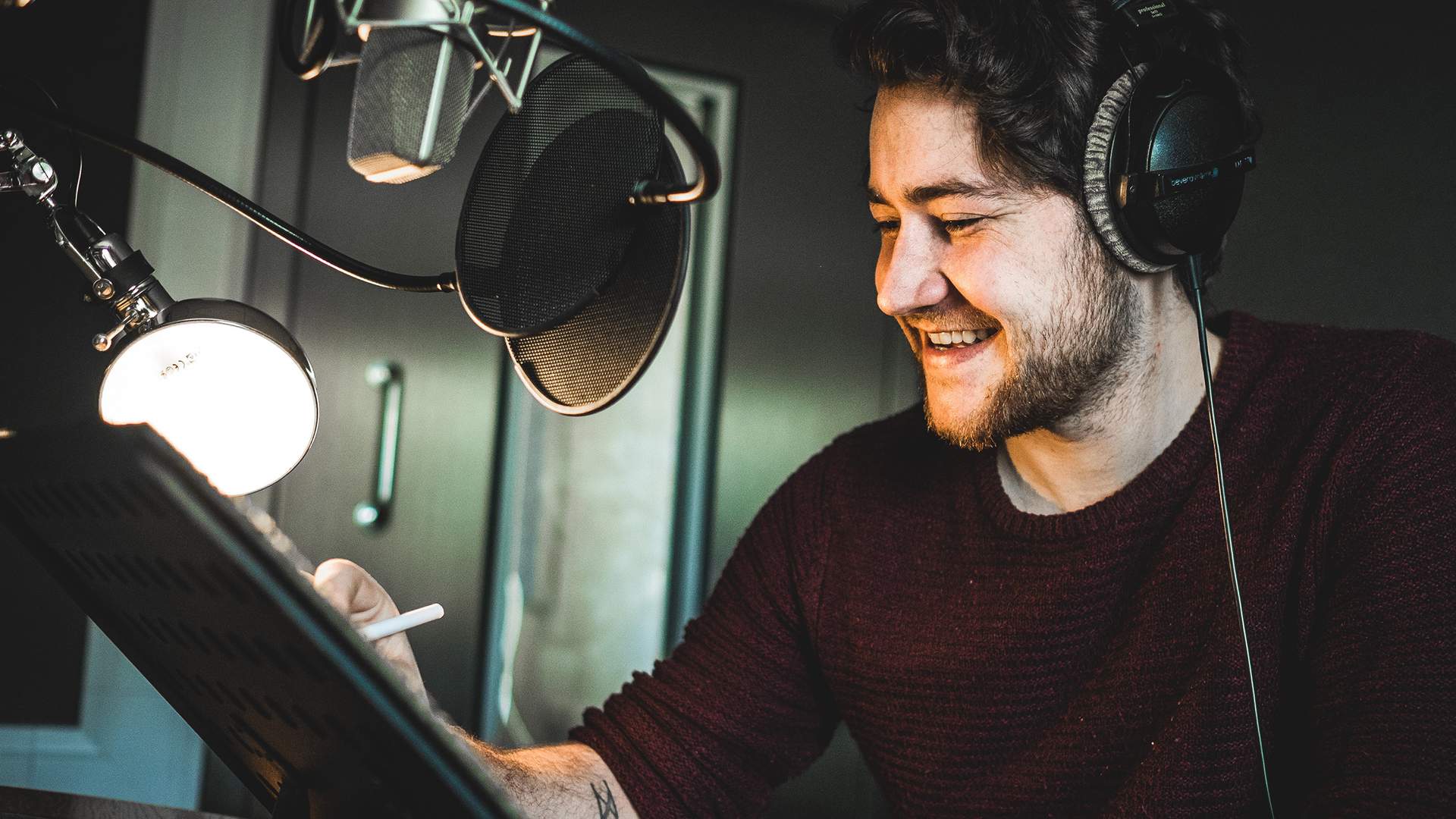 A man wearing headphones smiles while sitting in front of a microphone in a recording studio. He holds a pen and appears to be working or recording audio. The setting is dimly lit, with a lamp illuminating his workspace.