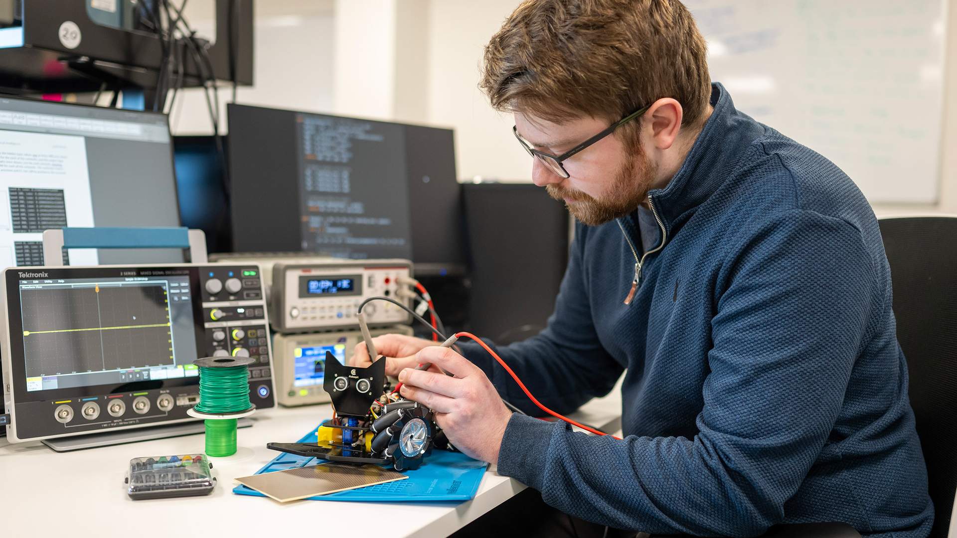 A person with glasses is seated at a desk in a lab, working on an electronic device with red wires. They are surrounded by equipment, including monitors and oscilloscopes. A whiteboard with writing is visible in the background.