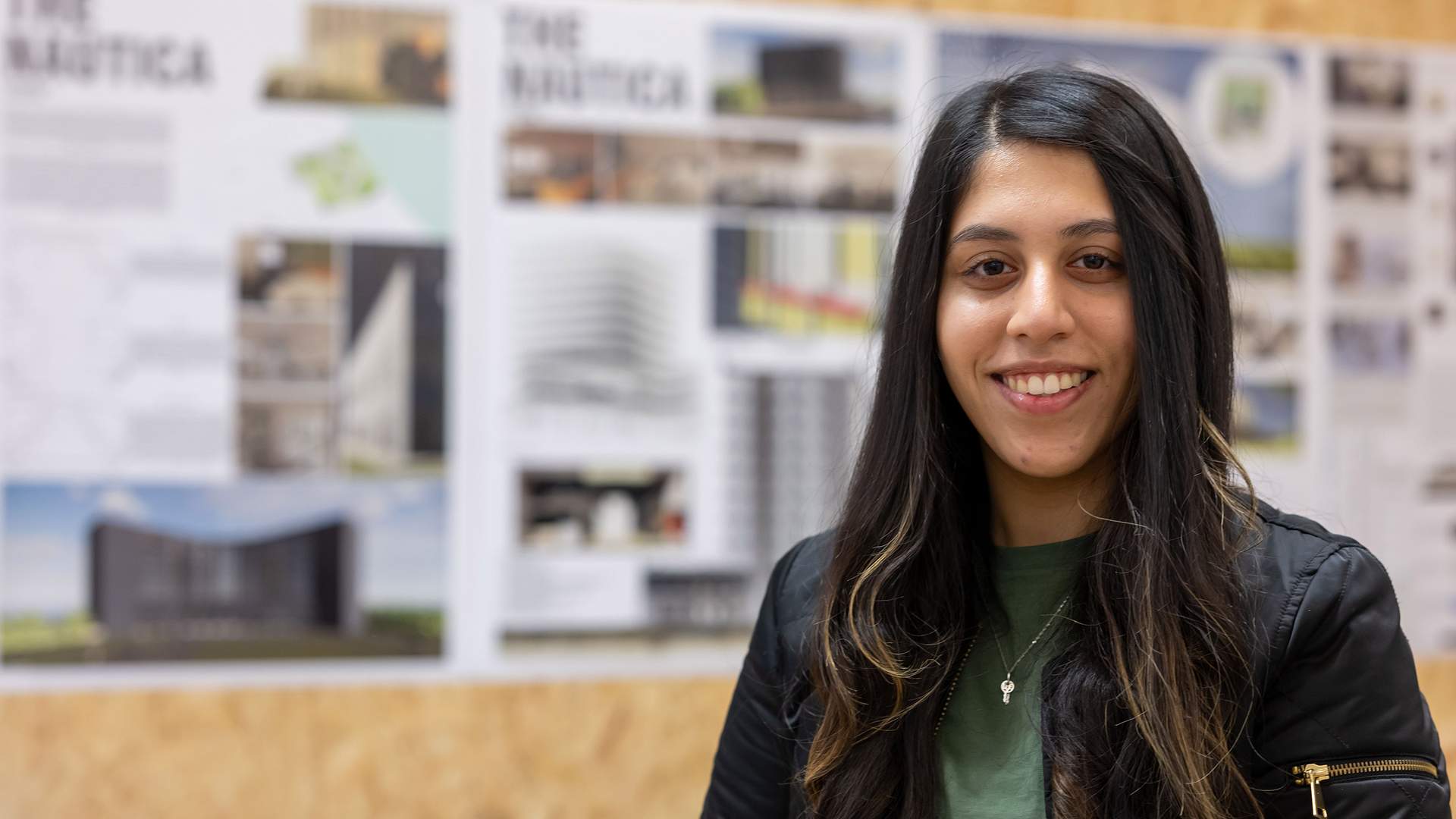 A person with long dark hair and a green shirt smiles at the camera. In the background, there are architectural design boards displayed on a cork wall.