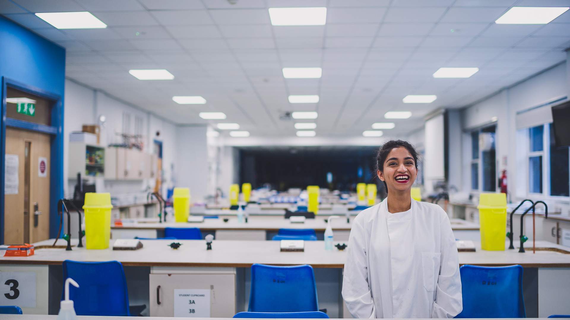 A woman with long hair stands confidently in a medical examination room, arms crossed. Behind her is a model skeleton, medical equipment, and a bed. She is wearing a dark cardigan over a white shirt and smiling.