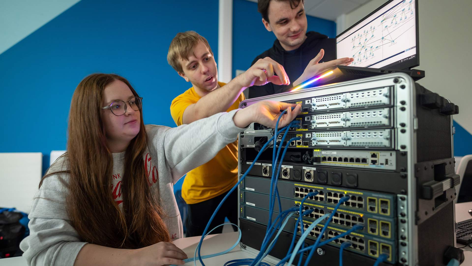 Three people work together on computer networking equipment. They are examining cables and a monitor displaying diagrams. The setting is a professional tech environment with a blue accent wall.