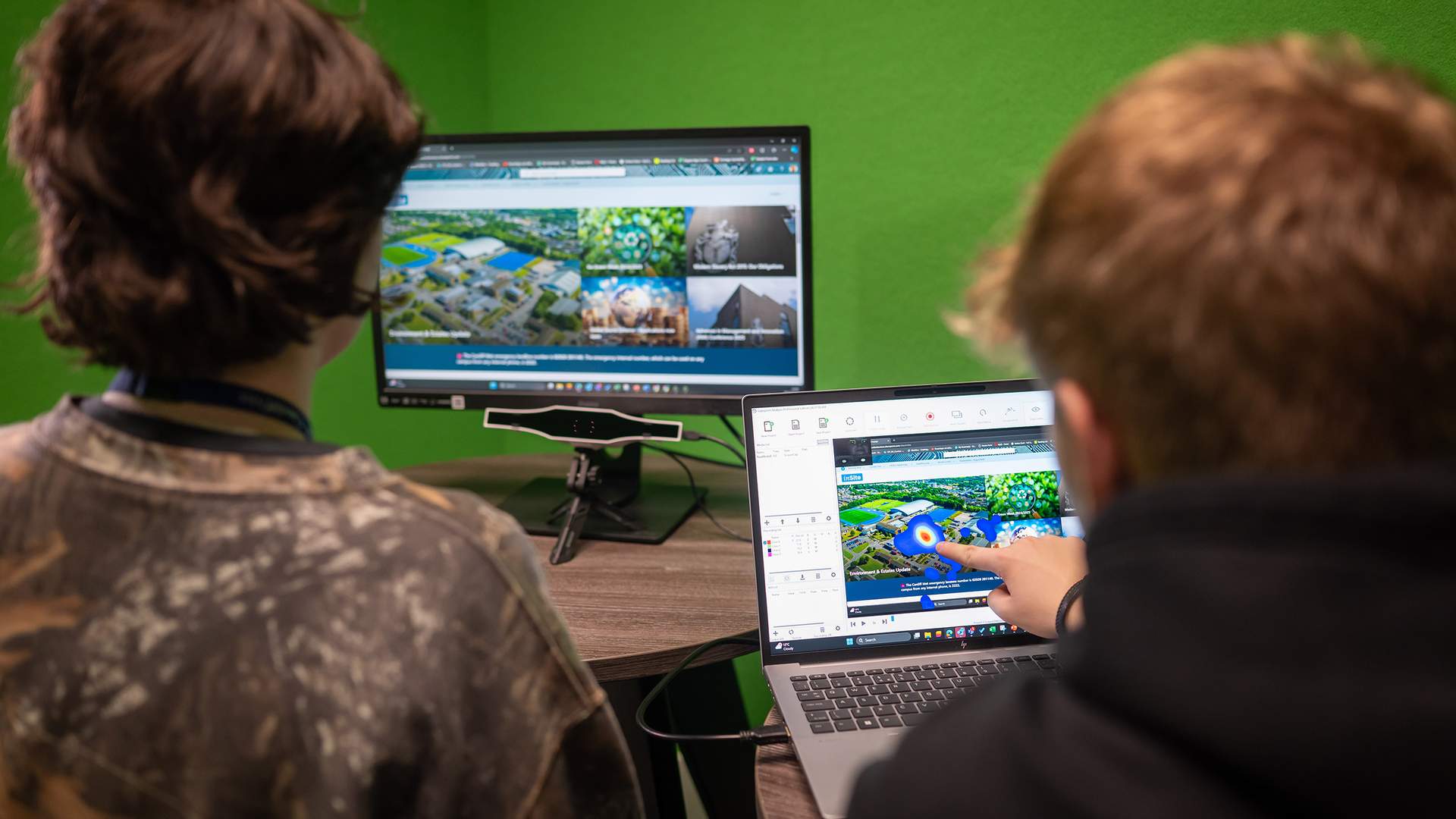 Two people are looking at computer screens in an office. One person points at a laptop displaying a design of a landscape, while a larger monitor shows similar images. The room has bright green walls.