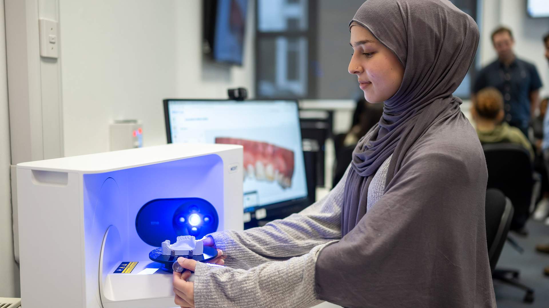 A person wearing a hijab operates a dental 3D scanning machine, placing a dental model inside. The setting appears to be an educational or professional environment, with a computer screen showing dental images in the background.