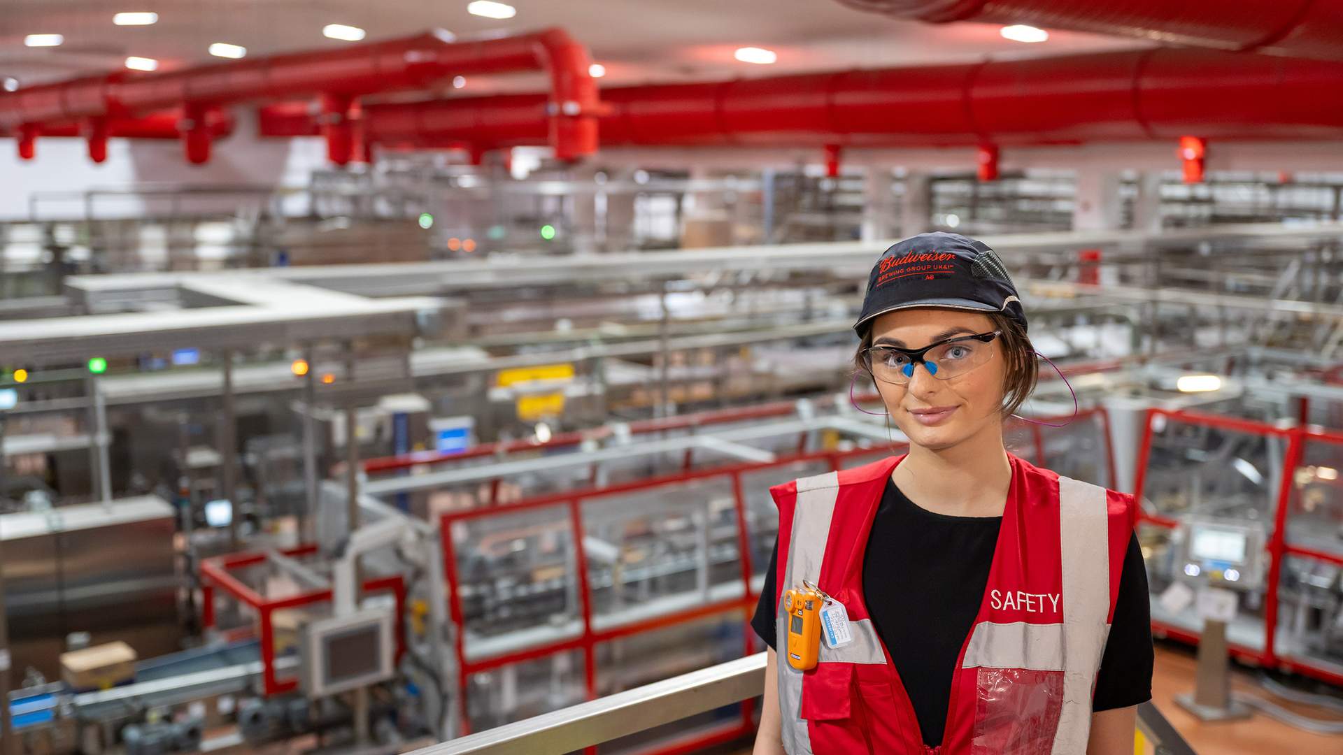 A person wearing safety gear and a cap stands in a large industrial facility with extensive machinery and red overhead pipes. The person is wearing a red vest labeled SAFETY and safety goggles, and looks toward the camera.