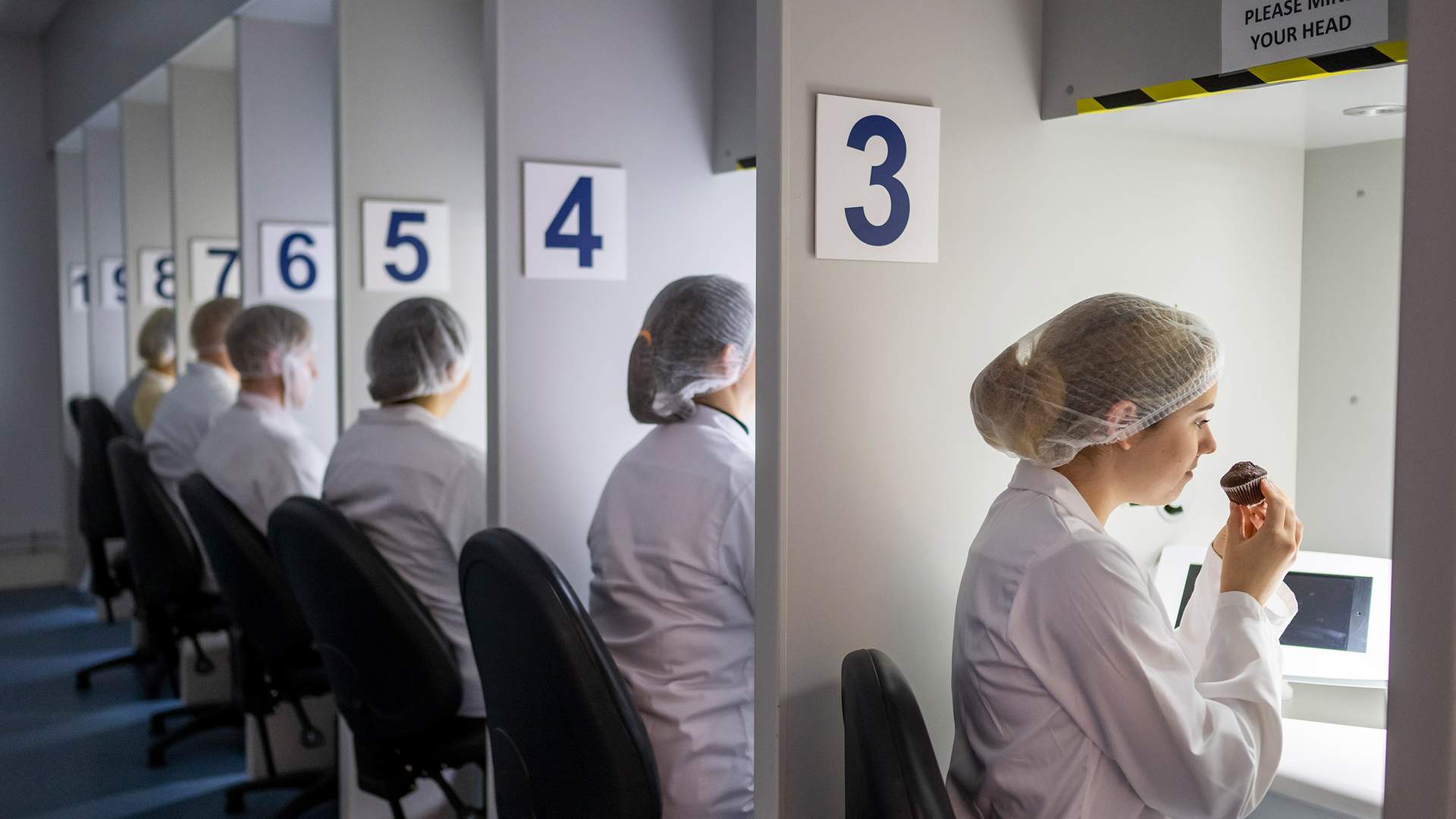 A row of people in white lab coats and hairnets sit at numbered stations. Each person is engaging in a sensory evaluation, with one person smelling or tasting from a small cup under a hooded light. The stations are numbered 3 to 8.