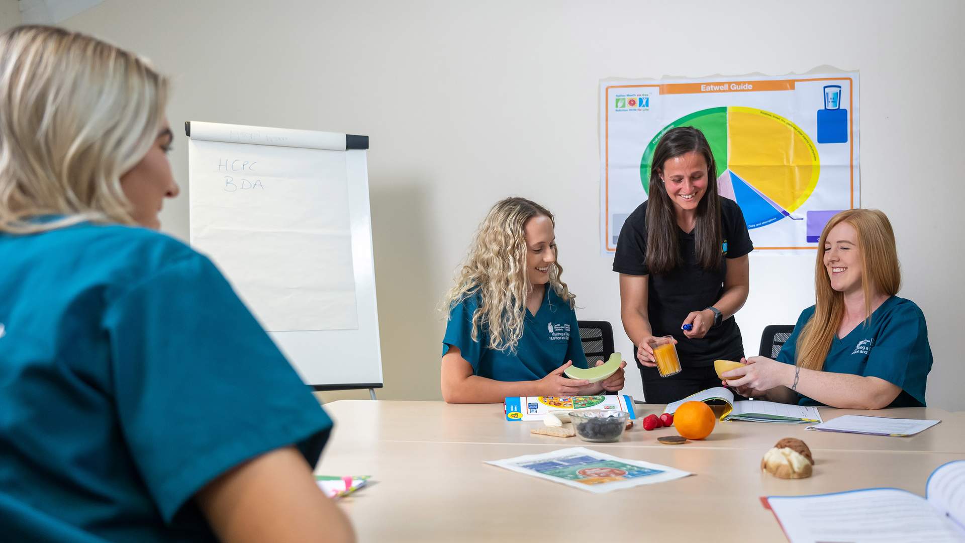 A group of women are seated at a table in a classroom setting, engaged in a discussion about nutrition. They have fruits on the table, and a pie chart on the wall is related to food groups. One woman is pouring a drink, while another peels a banana.