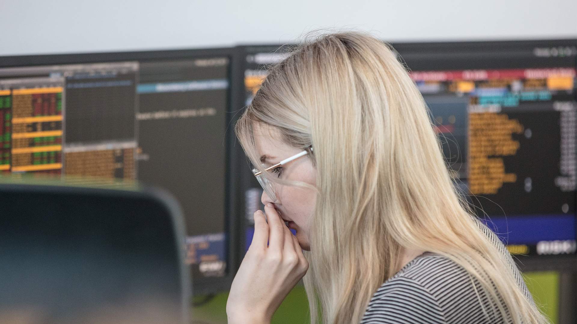 A woman with blonde hair and glasses is intently focused on monitors displaying complex data and graphs. She wears a striped shirt and appears to be in a busy office environment.