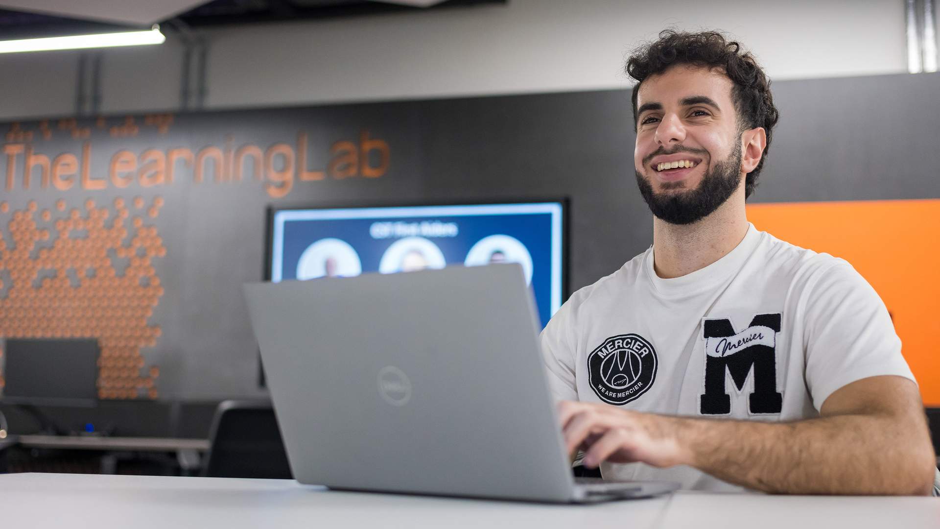 A person with a beard is smiling while using a laptop in a modern workspace. In the background, theres a screen and a wall with The Learning Lab text on it. They are wearing a white shirt with patches.