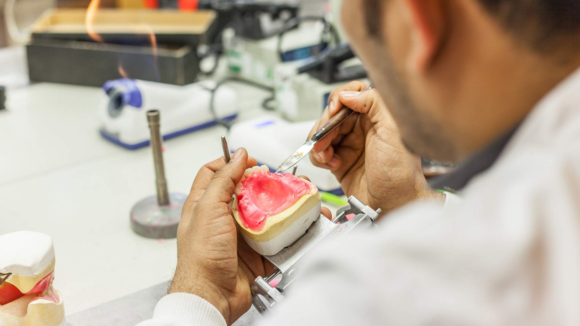 A dental technician works on a dental mold, meticulously shaping it with tools in a well-equipped lab. A small flame burner is visible on the table, suggesting a process that involves heat.
