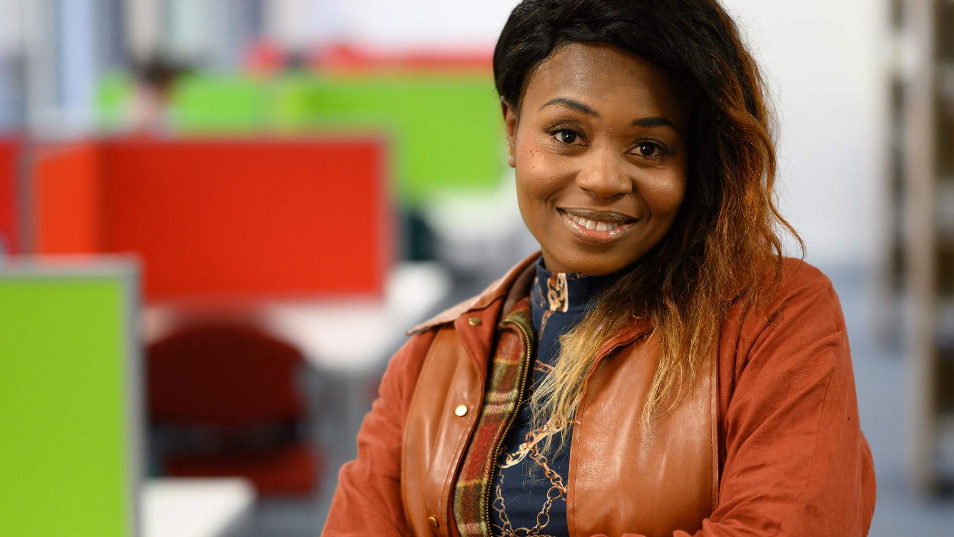 A woman with shoulder-length hair smiles warmly at the camera, standing in an office setting. She is wearing a brown jacket over a patterned top. The background features brightly colored partitioned workstations in red and green.