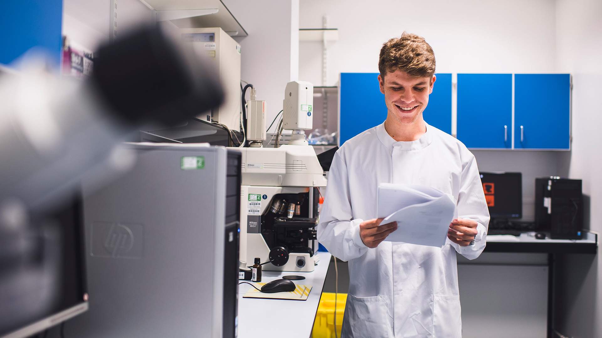A smiling person in a lab coat holds papers in a laboratory with microscopes and computers. Shelves with blue cabinets are in the background. The environment suggests a research or scientific setting.