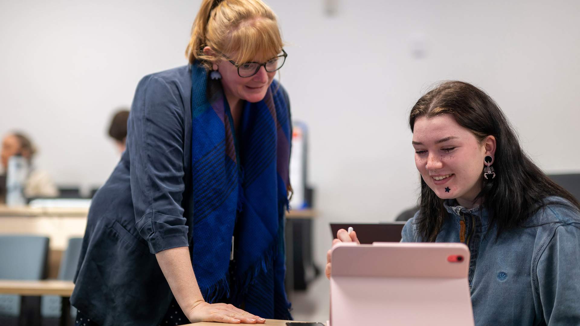 A teacher with glasses and a blue scarf leans over a students desk in a classroom. The student, smiling, is using a pink tablet. Other students appear in the blurred background.