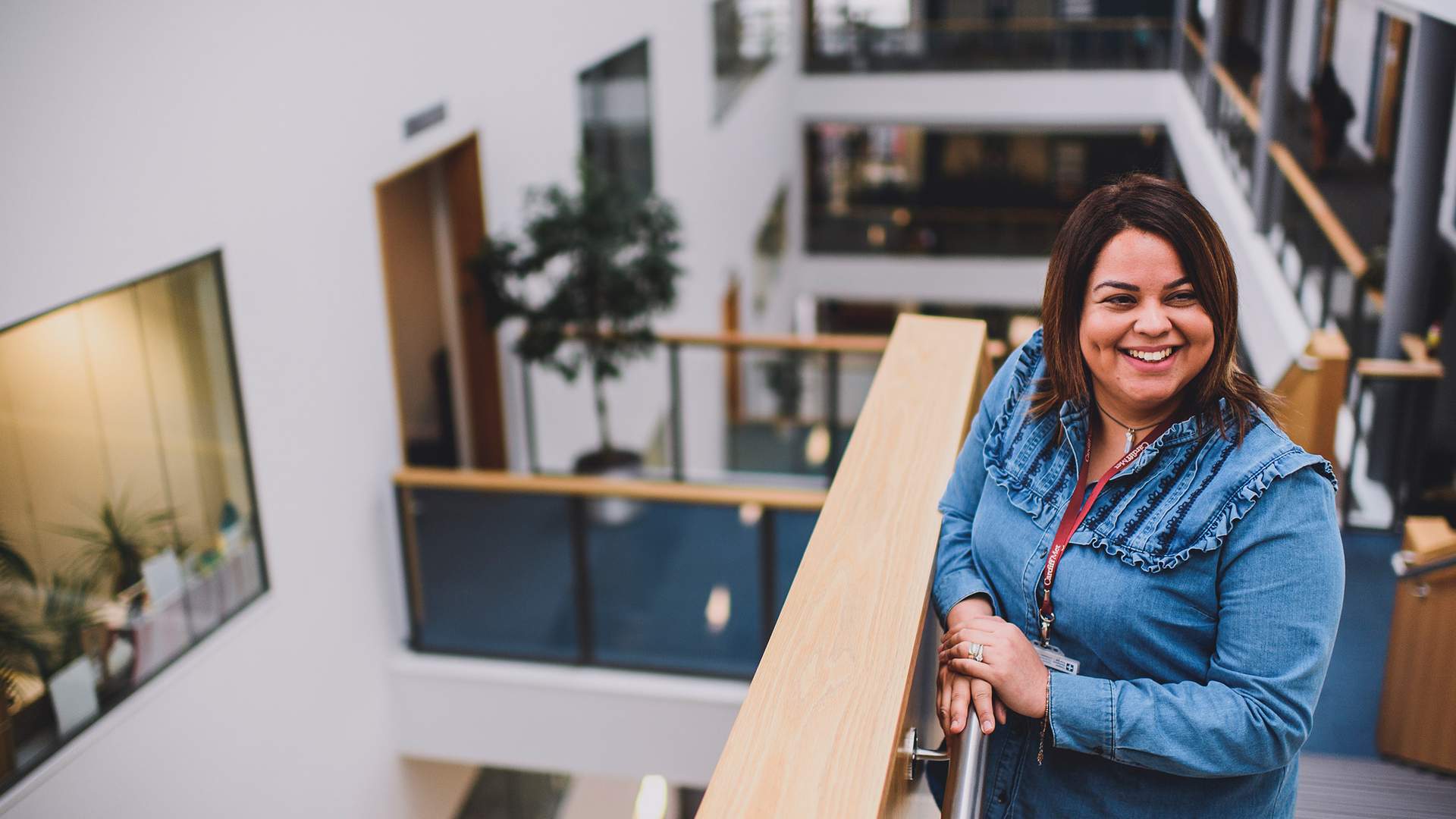 A woman smiles while leaning against a railing in a modern, multi-level office building. She is wearing a denim shirt and a lanyard. The background shows open hallways and glass barriers with indoor plants.